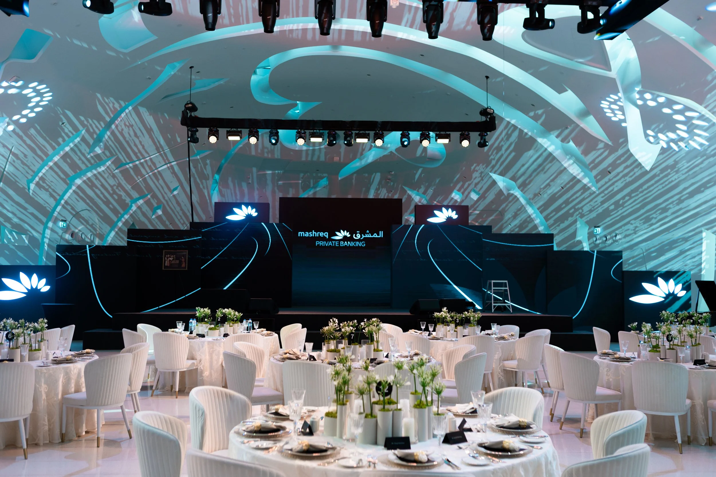 An elegant event space set up for a banquet with round tables covered in white tablecloths, decorated with white flowers and place settings. In the background, a modern stage with digital screens and lighting displays features the Mashreq bank logo, 
