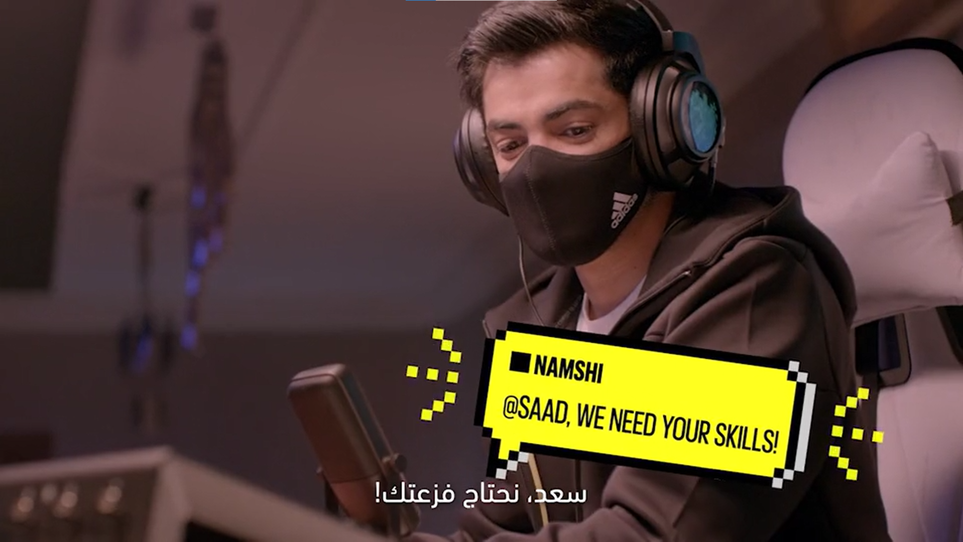 A young man with short dark hair wearing a black face mask and large headphones, sitting in an airplane seat, looking at his phone. There is a yellow pixelated graphic overlay with black text that reads '@SAAD, WE NEED YOUR SKILLS!' and Arabic text a