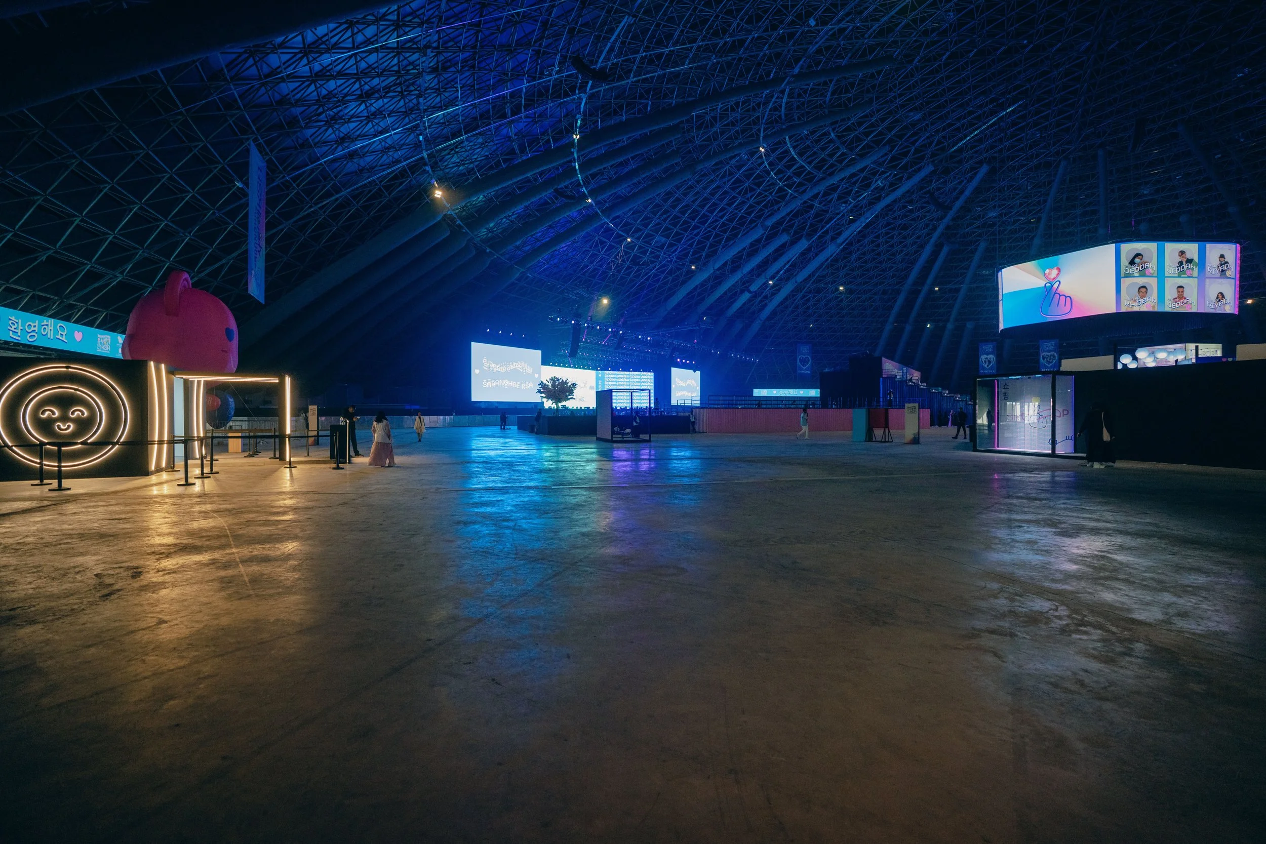 Empty indoor arena or exhibition hall with large LED screens, illuminated displays, and a few people walking around.