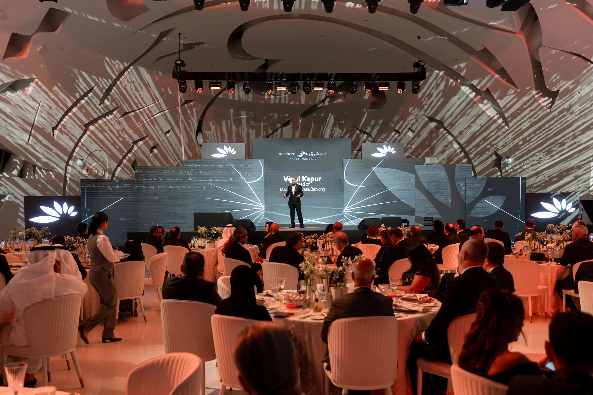 A speaker on stage at a formal event or conference with an audience seated at round tables. The stage background features digital graphics and the logo of Mashreq Bank, with a person standing and presenting.