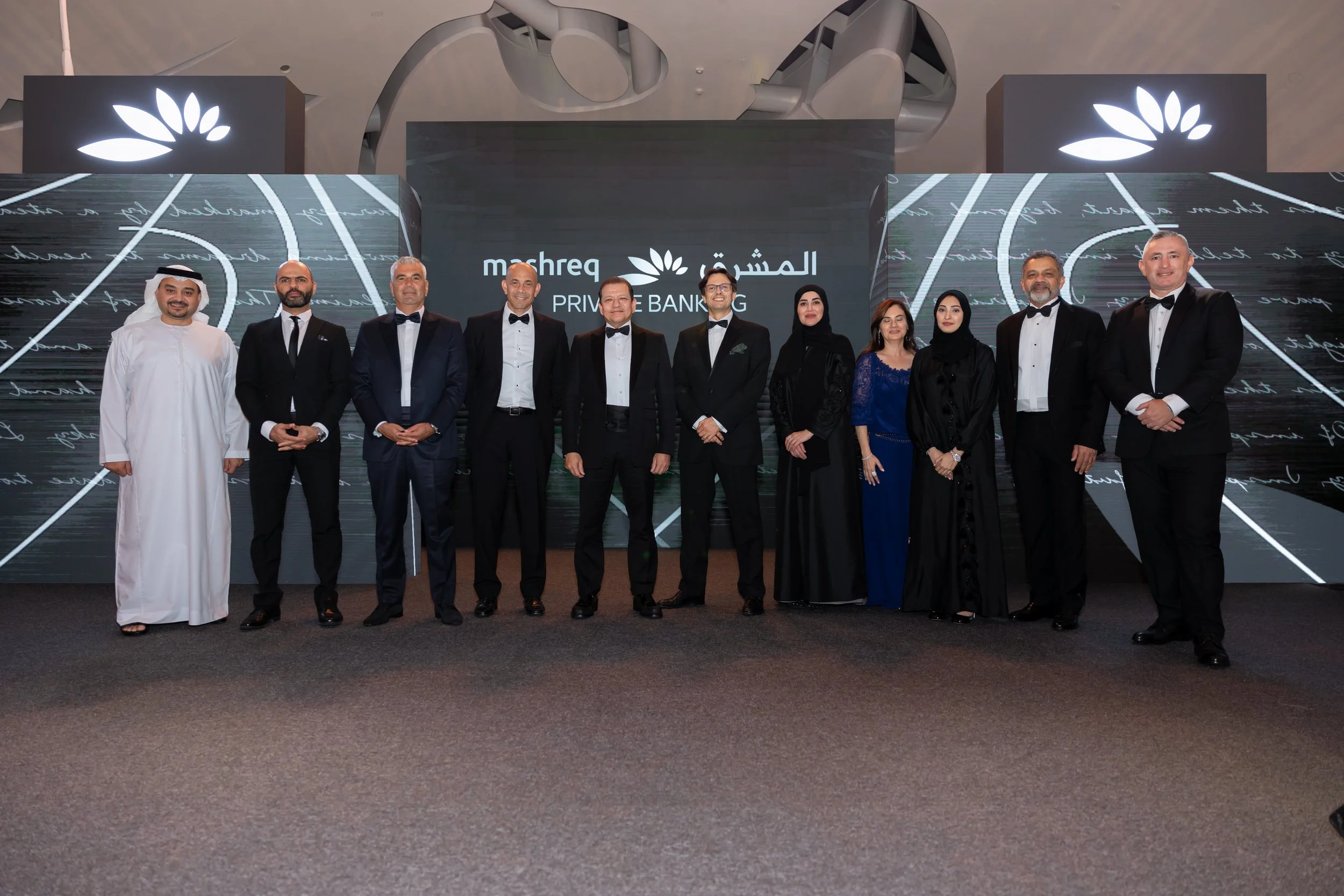 A group of twelve people dressed in formal attire, including tuxedos and traditional Middle Eastern clothing, standing on a stage with a digital screen behind them displaying Arabic text and the words 'Mashreq Private Banking' in English.