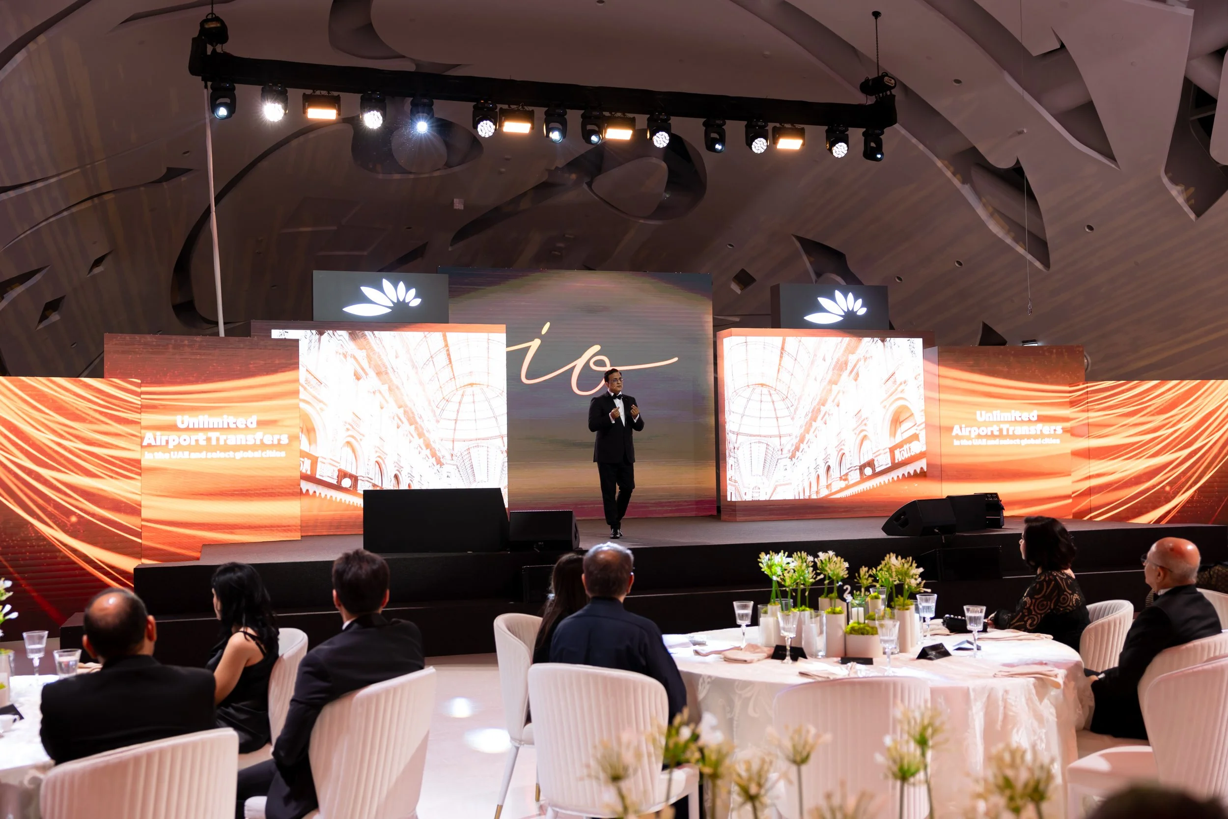 A speaker in a suit on stage at a banquet or conference, presenting to an audience seated at round tables with white tablecloths and floral centerpieces, stage with large digital screens displaying the text 'Unlimited Airport Transfers in the UAE and