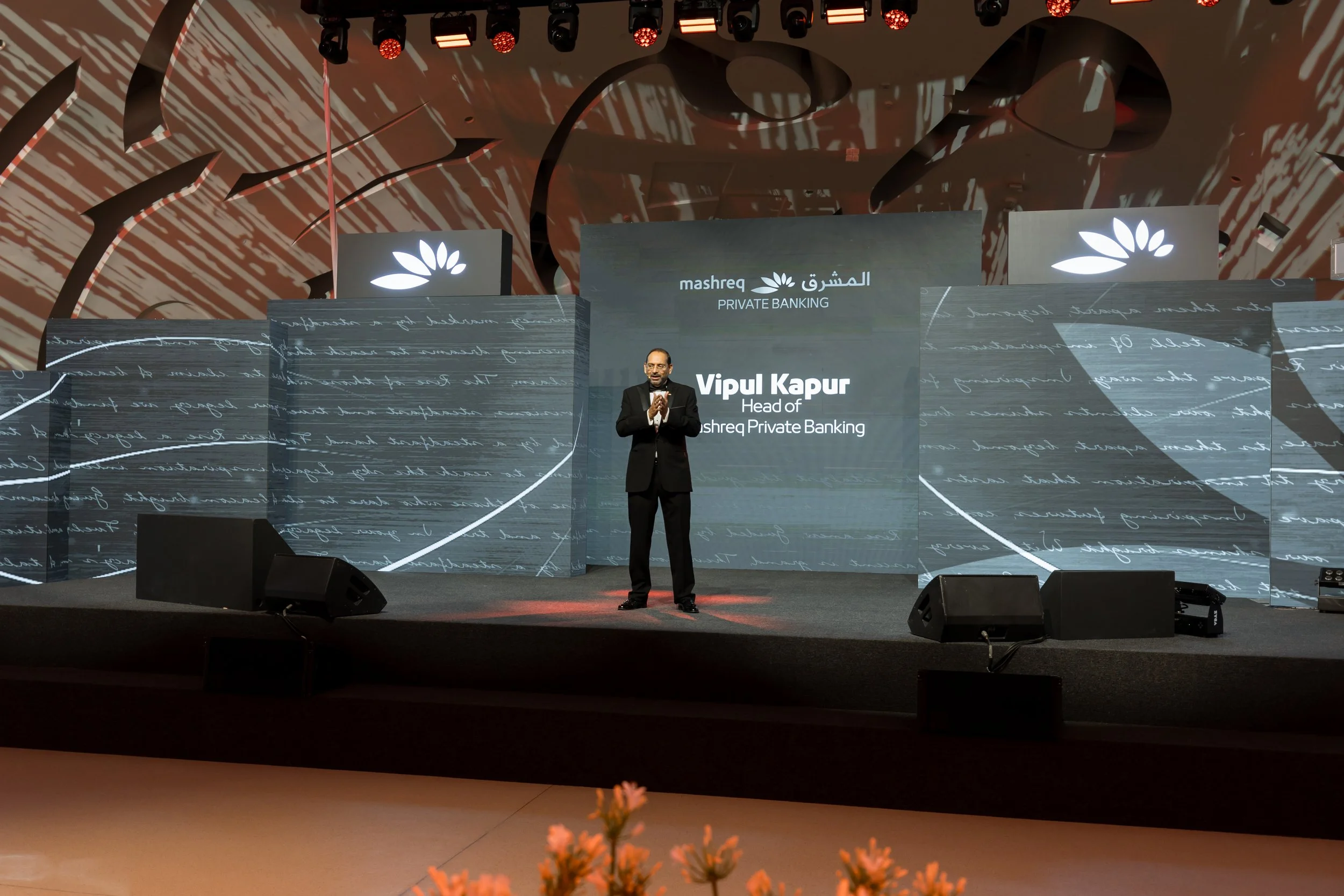 Vipul Kapur speaking on stage at a corporate event organized by Mashreq Private Banking, with a large screen behind him displaying his name and title, and abstract background visuals projected on the stage backdrop.