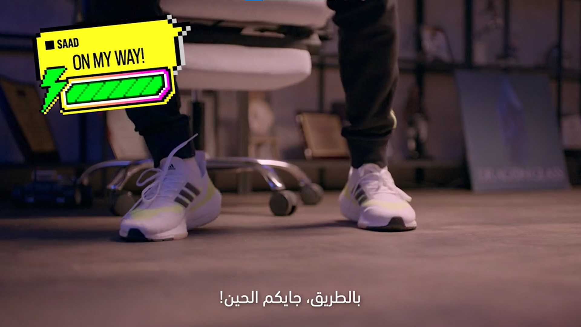 Close-up of a person's feet in white sneakers with yellow and black stripes, standing on a wooden floor. There is a colorful graphic overlay with the text 'SAAD ON MY WAY!' and a battery icon. The background shows a desk, some chairs, and a poster, w