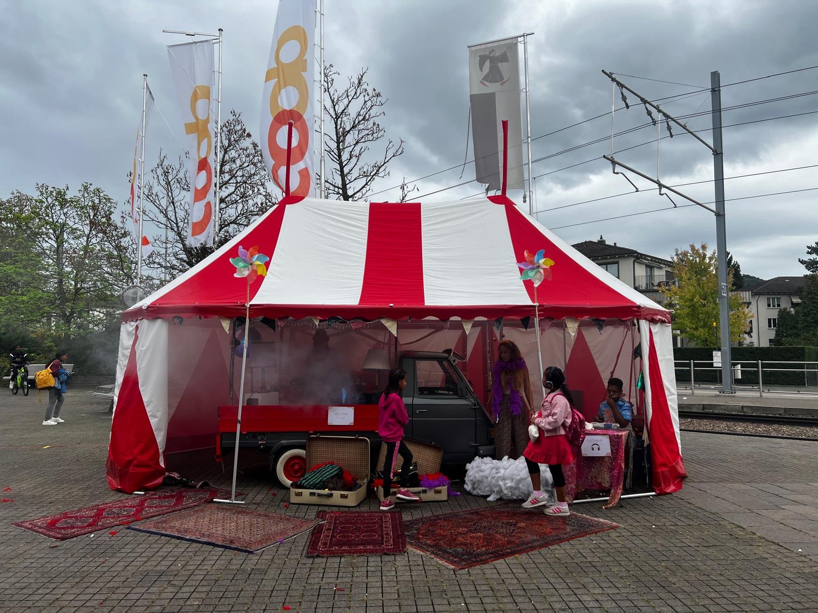 Ein buntes Zirkuszelt mit Menschen und Kindern davor, mittelgroße Stadtarchitektur im Hintergrund, Wolkenhimmel, auf dem Boden Teppiche und alte Koffer, im Stadtverkehr.
