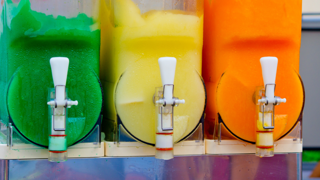 Mehrere Getränke in großen transparenten Tanks mit Ausgüssen, grün, gelb und orange gefärbt, wahrscheinlich Fruchtsaft-Getränke.