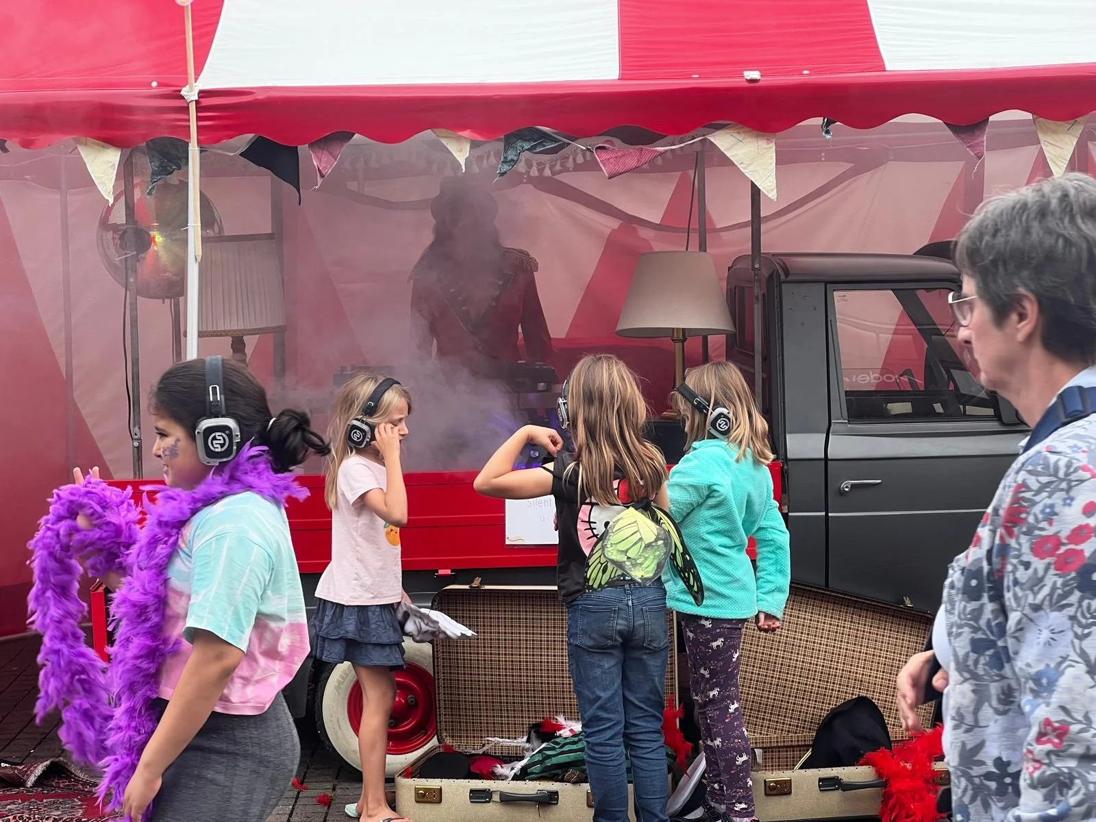 Mehrere Kinder tragen Kopfhörer und Kostüme, stehen vor einem roten Verkaufswagen mit einem DJ im Inneren, der Musik auflegt. Es ist eine festliche Atmosphäre.