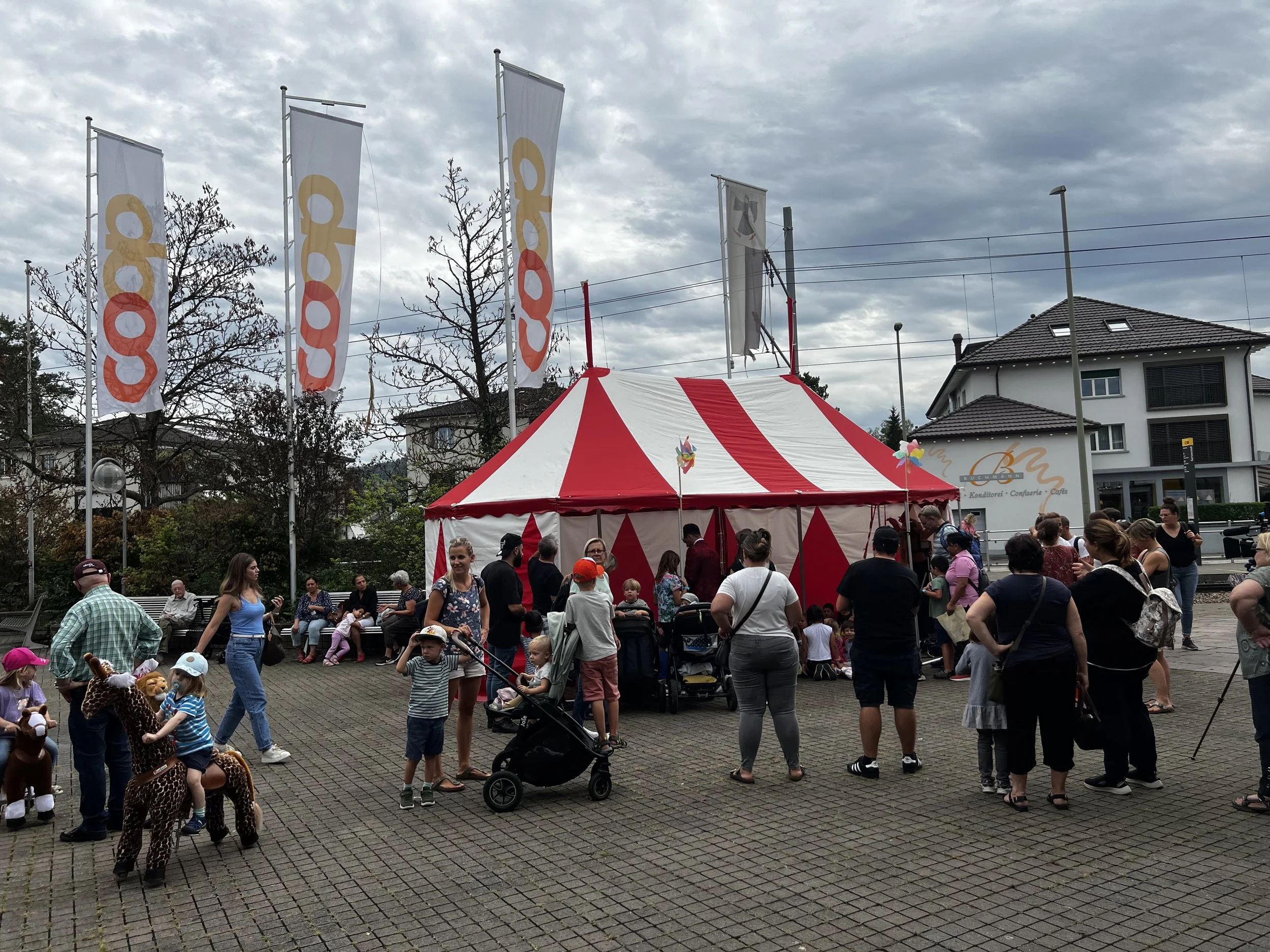 Ein Zirkuszelt mit rotem und weißem Streifen, umgeben von Menschen, darunter Kinder und Erwachsene, einige in Tierkostümen, auf einer gepflasterten Fläche an einem bewölkten Tag.