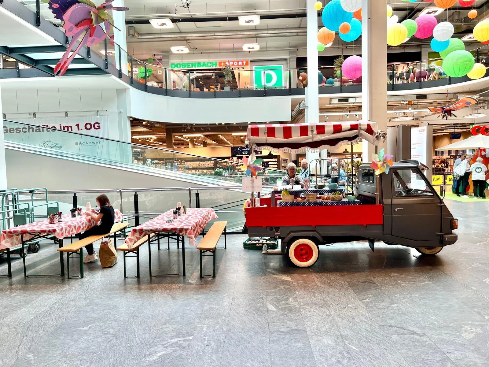 Ein kleiner Verkaufswagen in Form eines Dreirads im Einkaufszentrum mit Tischen und Sitzbänken vor dem Wagen. Kinder sitzen an einem der Tische und kaufen. Das Einkaufszentrum ist bunt dekoriert mit Papierlampen und kann auch eine Familie beim Einkau