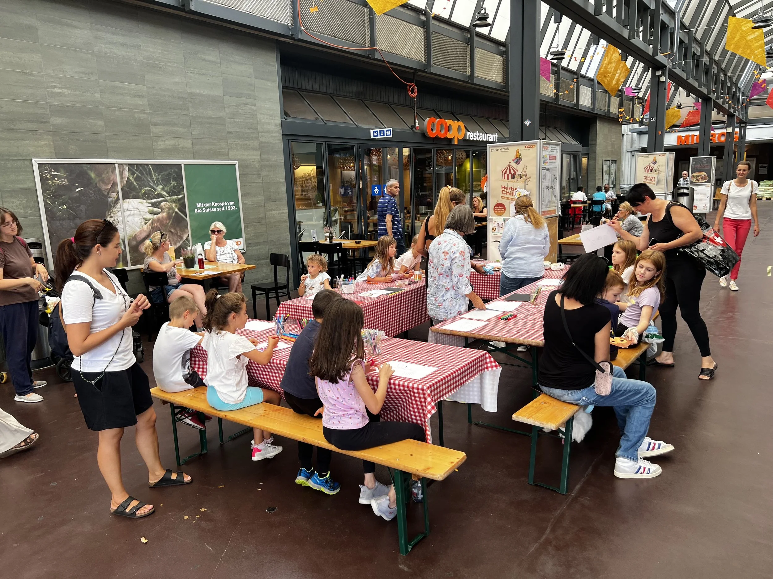 Menschen sitzen und stehen bei einem Kinderaktivitäts-Event mit Tischen, auf denen Papier und Stifte liegen, in einem überdachten Einkaufszentrum vor einem Coop-Restaurant.