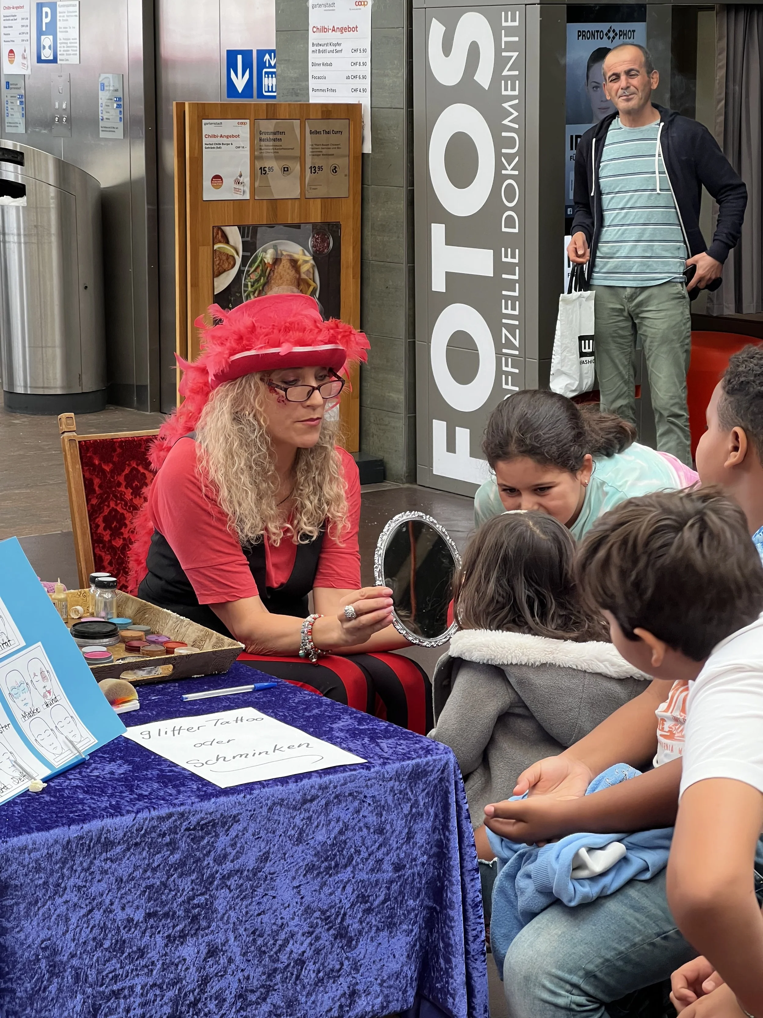 Eine Frau in einem roten Hut mit Federn zeigt den Kindern einen handgefertigten Spiegel bei einer Kinderaktivität, um Schminken oder Tattoos aus Glas oder Glitzer zu machen, am Tisch mit einem blauen Samtkleid und einem Schild auf dem Tisch.