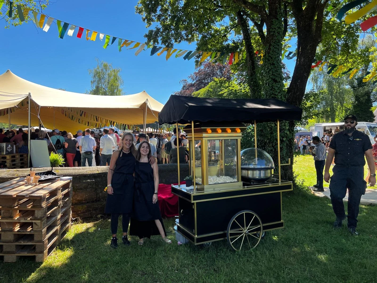 Zwei Frauen stehen mit einem Popcornstand bei einem Open-Air-Festival, eingerahmt von bunten Fahnen unter einem großen Baum, mit mehreren Zelten und Menschen im Hintergrund.