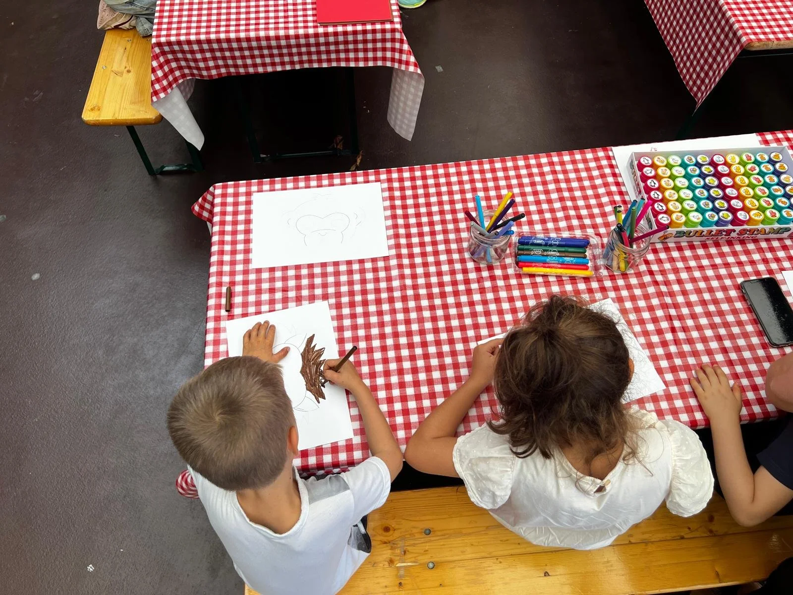 Zwei Kinder, ein Junge und ein Mädchen, sitzen an einem Tisch mit rotem und weißem kariertem Tuch und machen Bastelarbeiten. Der Junge zeichnet auf Papier, das Mädchen klebt ein Blatt mit braunem Papier.