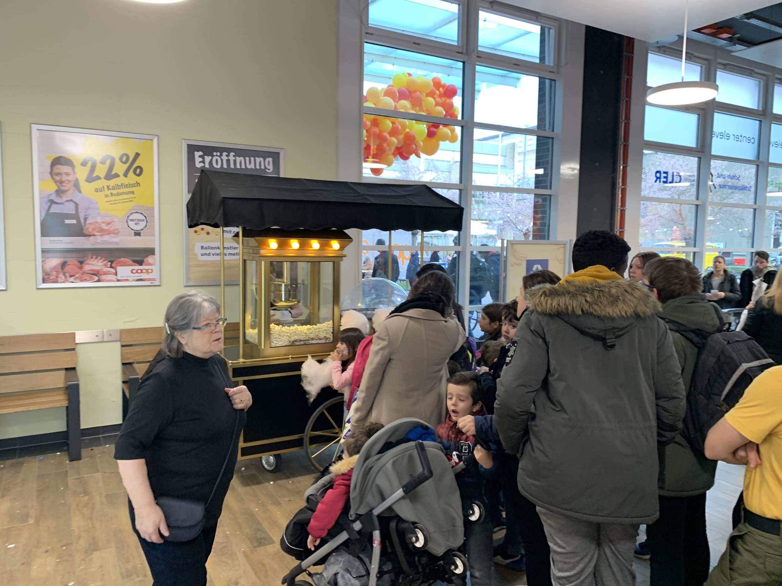 Menschenmenge an einem Popcorn-Stand in einem Supermarkt, im Hintergrund bunte Ballons, Poster an der Wand, große Fenster, natürliche Lichtquelle.