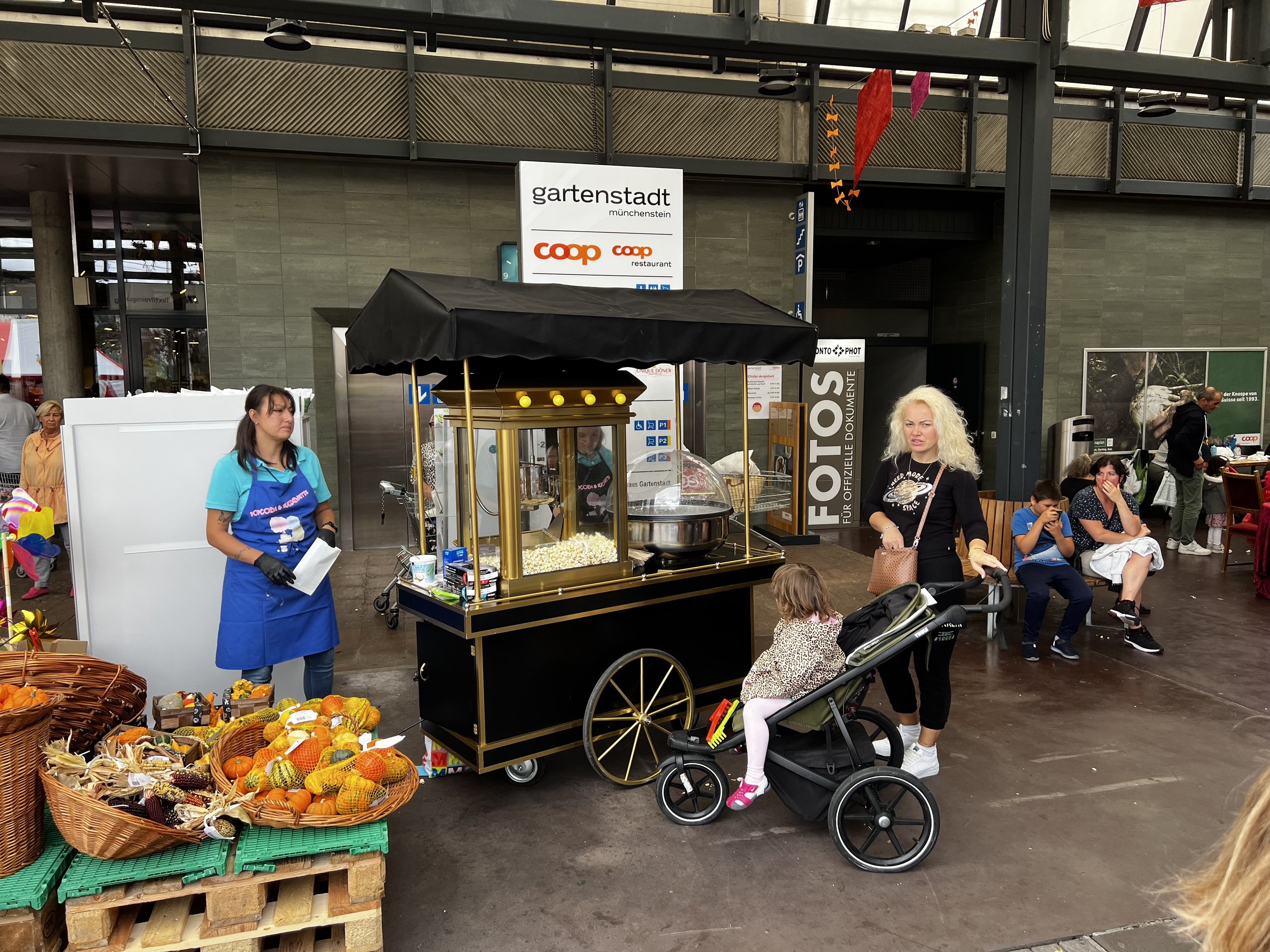 Menschen an einem Popcorn-Stand auf einem Markt in einer großen Halle, umgeben von Sitzgruppen, mit mehreren Kindern und Erwachsenen, im Hintergrund Café und Sitzplätze.
