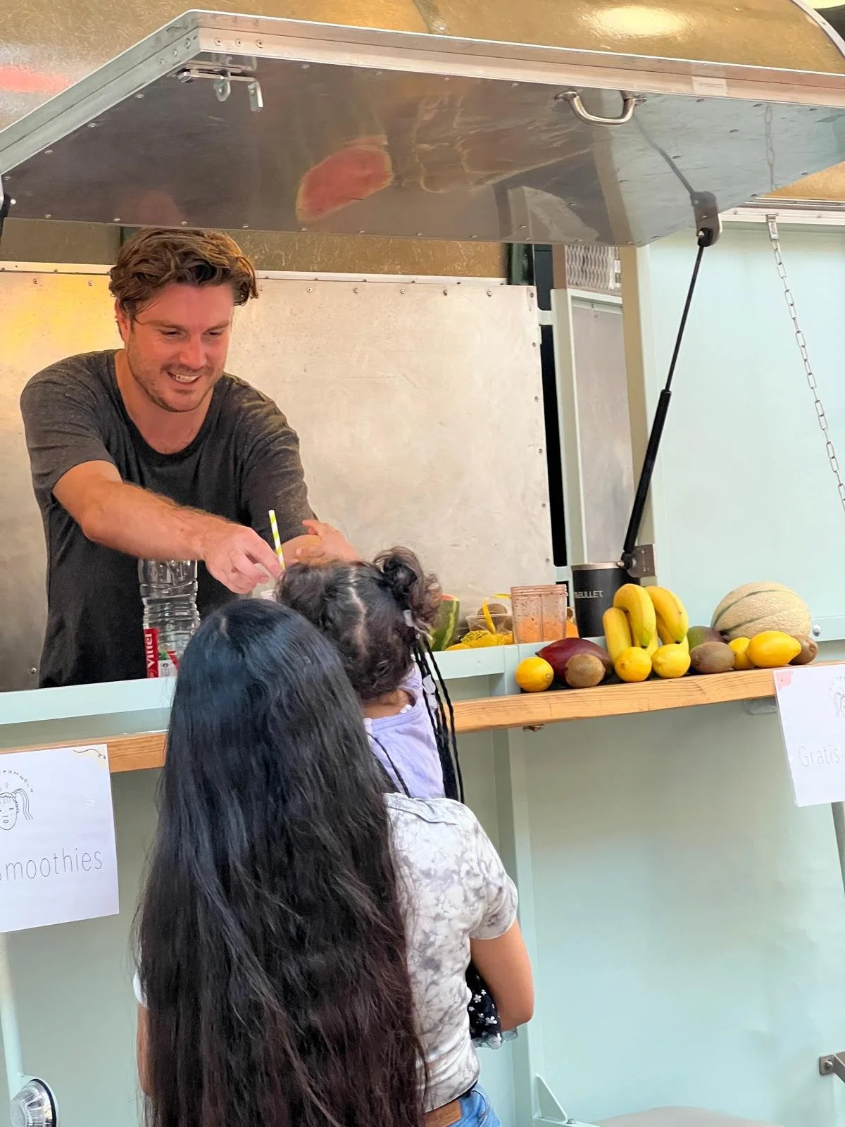 Ein Mann verkauft Früchte und Getränke an zwei Frauen an einem Fruchtstand, auf dem Bananen, Melonen und andere Früchte liegen.