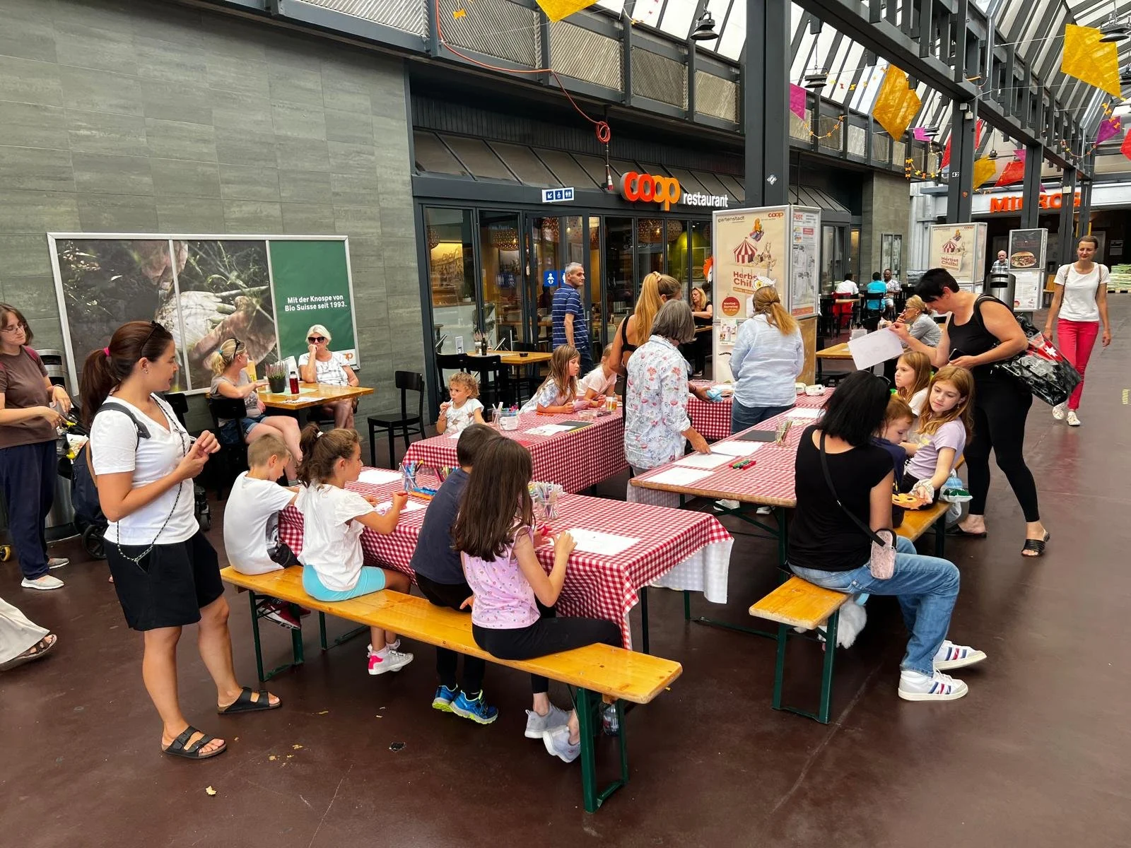 Familien beim Essen an einem Outdoor-Tisch mit rot-weiß kariertem Tischdecken vor einem Coop-Supermarkt.