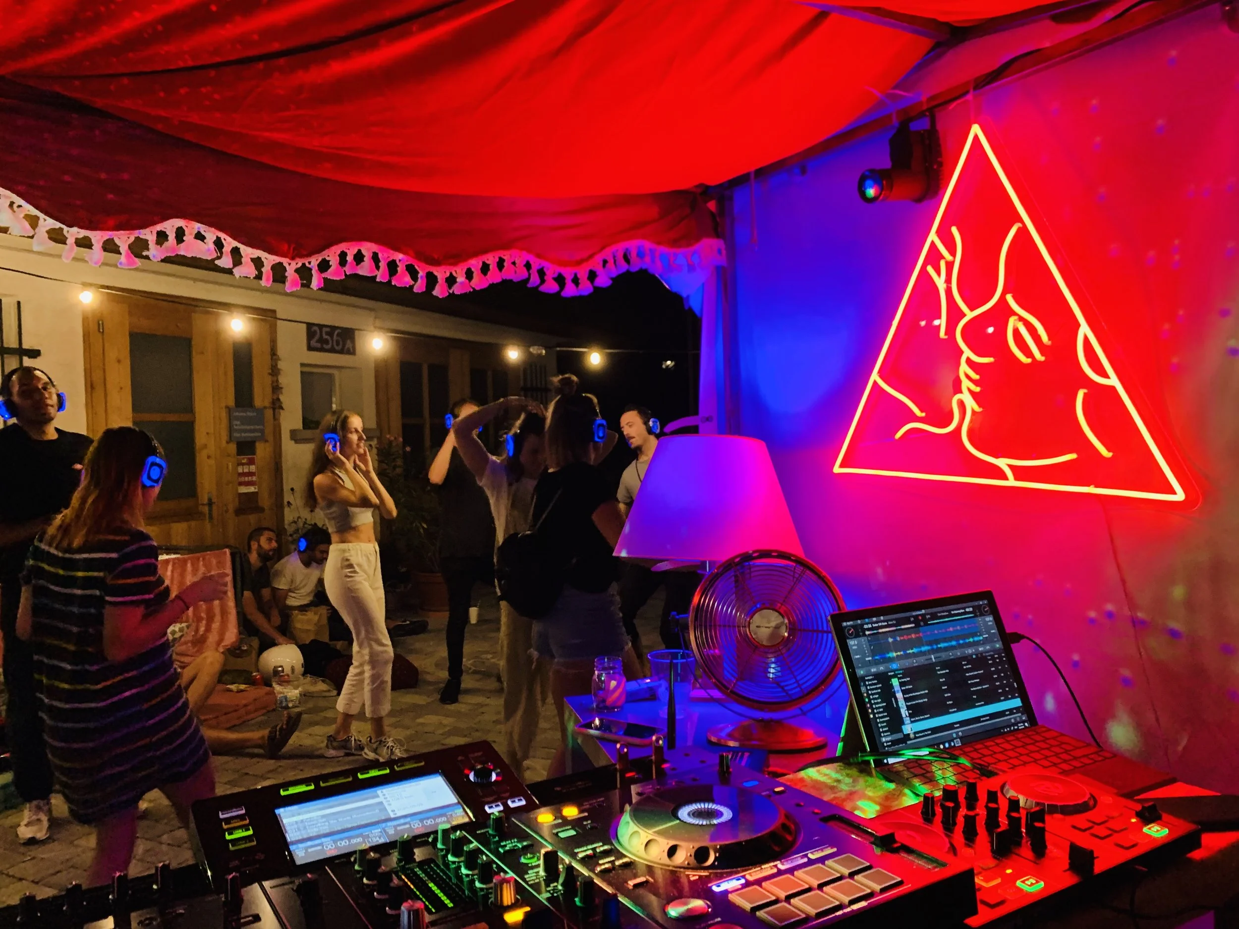 Unterhaltung in einer Party- oder Club-Atmosphäre mit Menschen, die mit Kopfhörern tanzen, und einer DJ-Station mit Mixer und Laptop, vor einer Neon-Signatur eines Liebespaares.
