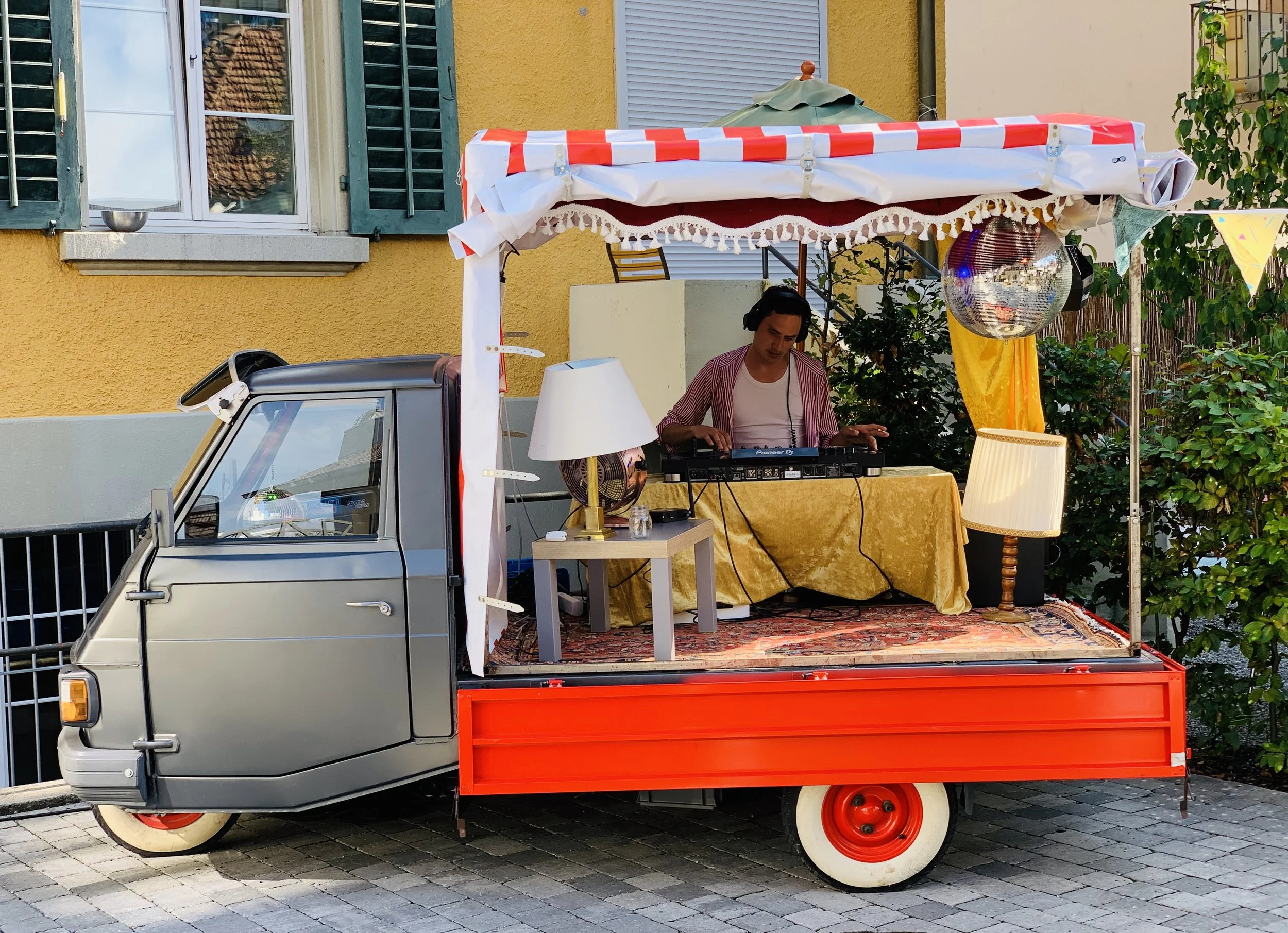 Ein kleiner aufgemotzter dreirädriger Lieferwagen mit einem DJ, der Musik macht, dekoriert mit Lampen, einem Spiegelball und bunten Fahnen, steht auf einer gepflasterten Straße vor einem gelben Haus mit grünen Fensterläden.