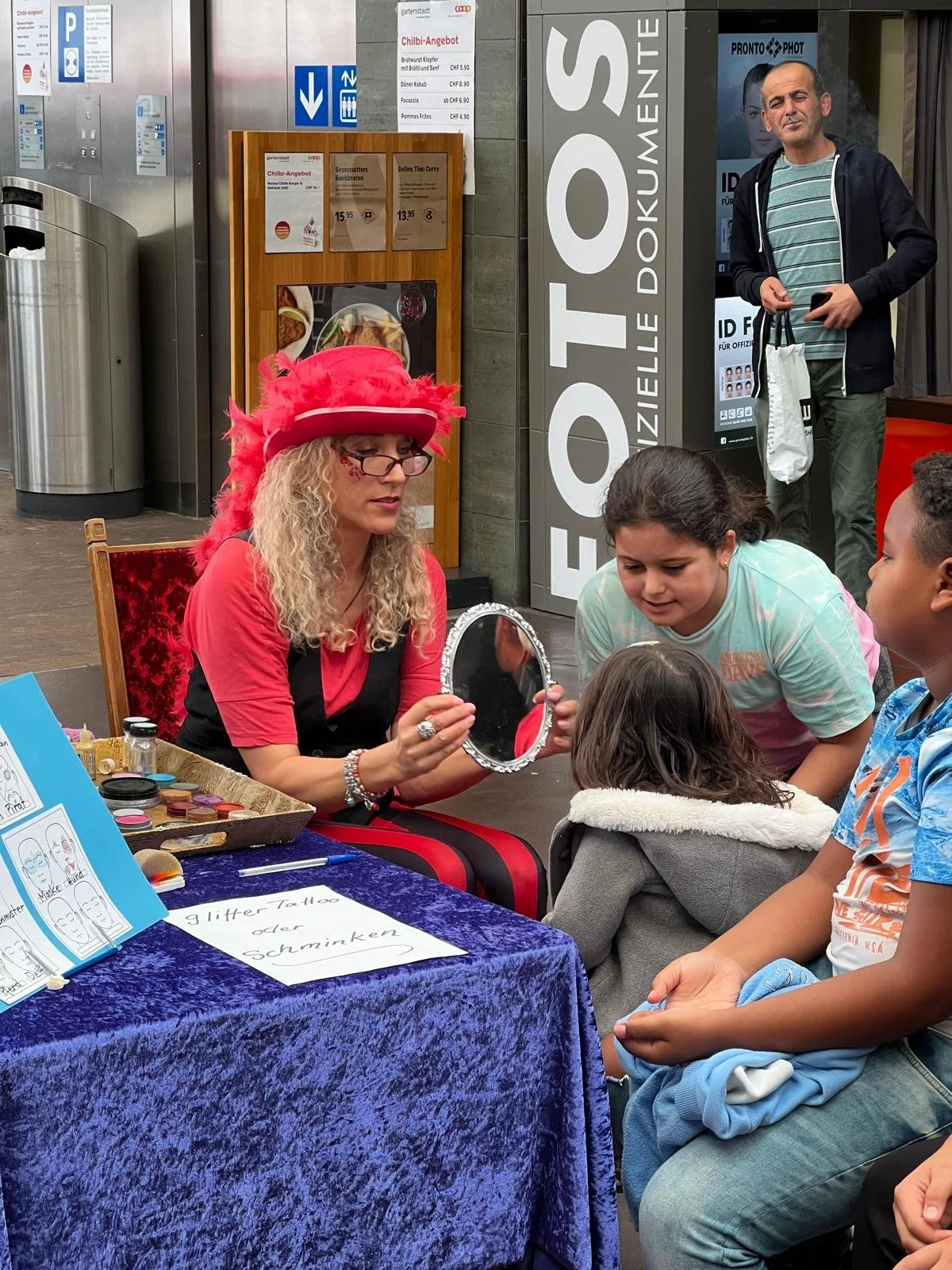Eine Frau in einem roten Hut und Brille macht Make-up vor einem Spiegel für Kinder. Es gibt eine Tafel mit Angeboten für Glitzer-Tattoos oder Schminken, und einige Kinder beobachten sie.