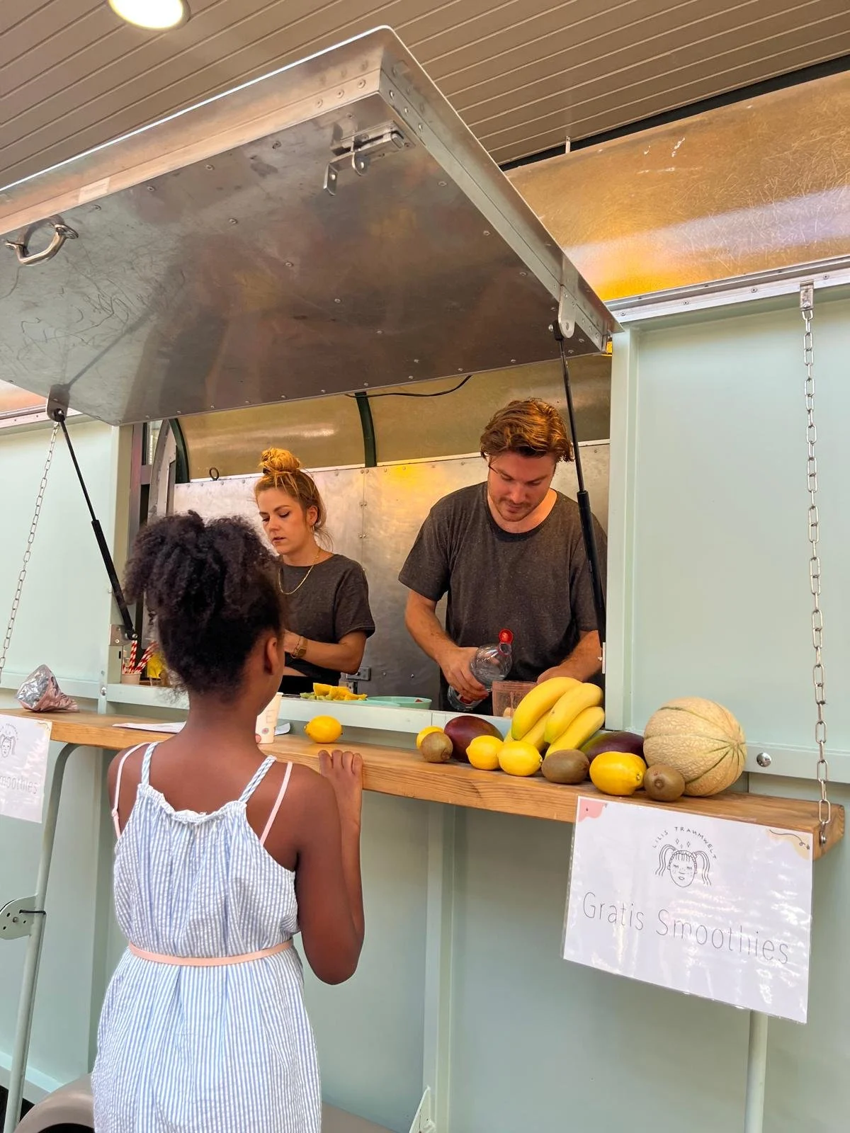 Zwei Menschen hinter einem Stehverkaufstisch mit Früchten, eine junge Mädchen kauft, die Früchte sind Bananen, Kiwis, Melone, Mango, Passionsfrüchte, Zitronen und Limetten. Ein Schild liest 'Gratis Smoothies'.