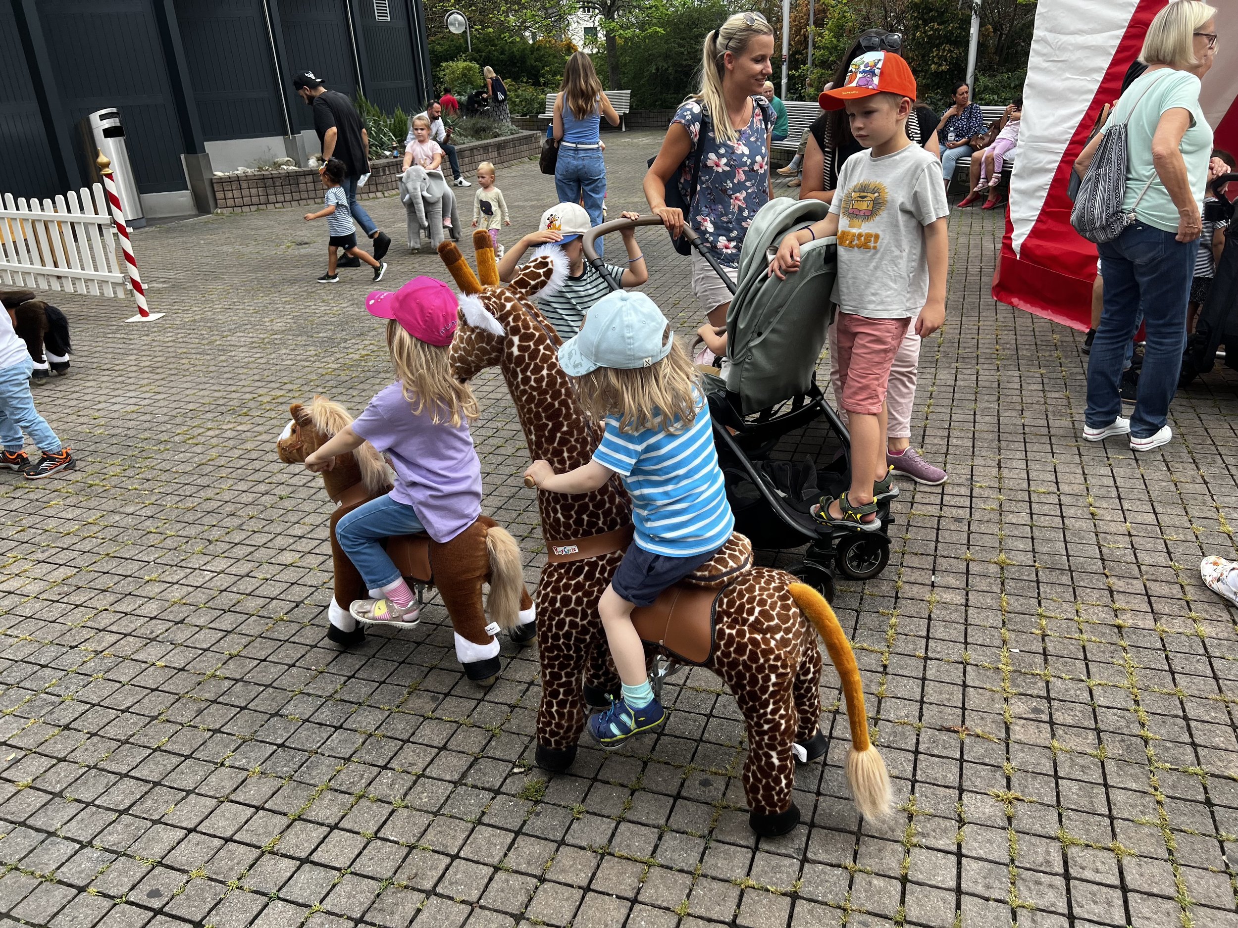 Kinder in Giraffen- und Pferdekostümen reiten auf Spielzeugpferden auf einem gepflasterten Spielplatz, während Erwachsene und andere Kinder im Hintergrund unterwegs sind.