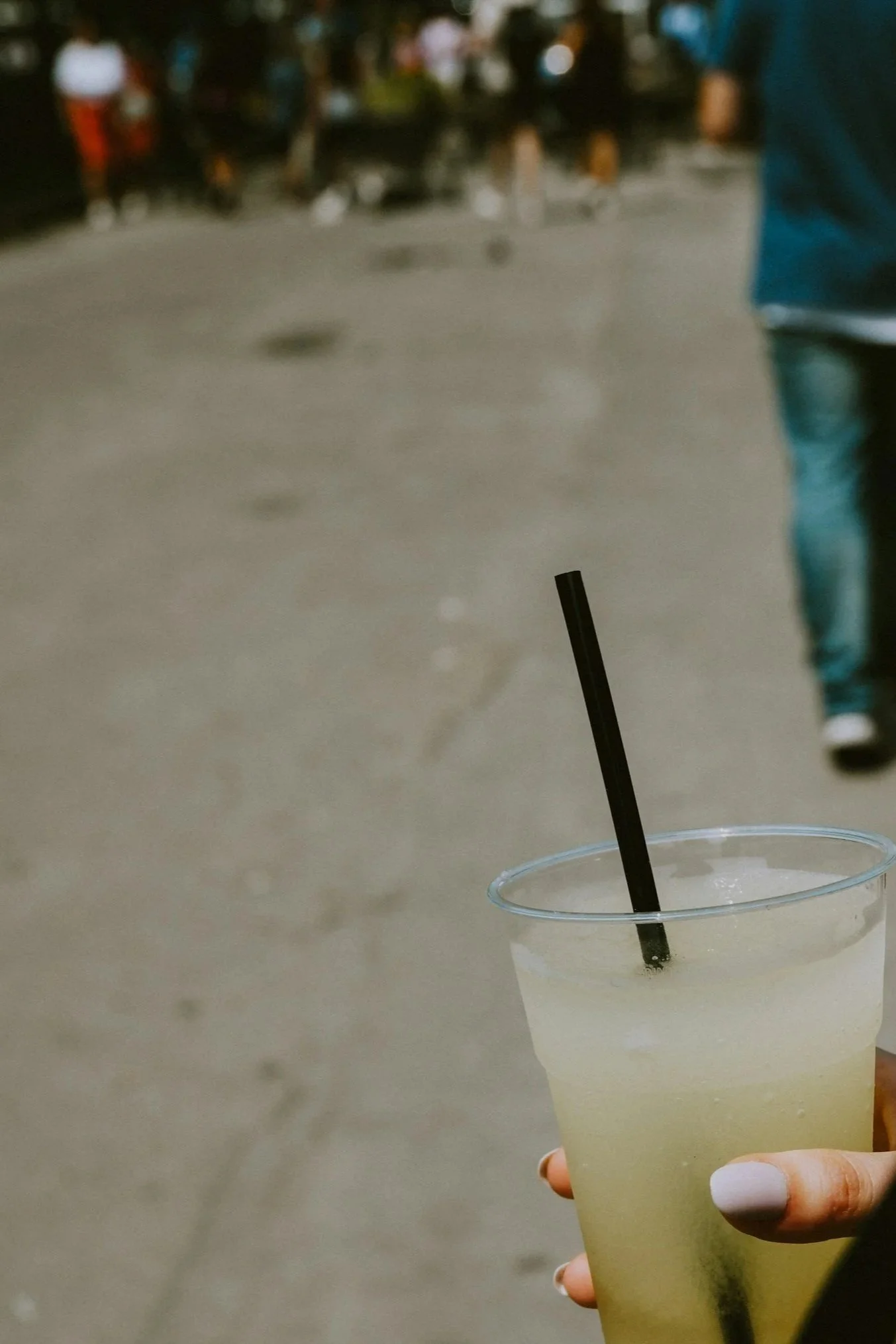 Eine Hand hält ein gut gefülltes transparentes Plastikglas mit einem Getränk und einem schwarzen Strohhalm, im Hintergrund sieht man eine belebte Stadtstraße bei Nacht.