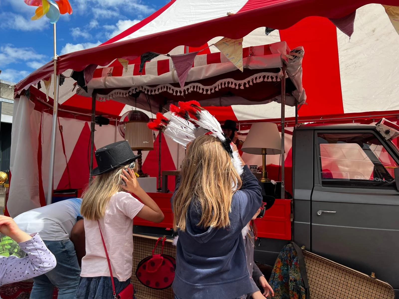 Kinder mit Hüten und ein Zirkuszelt mit roten und weißen Streifen, bunter Himmel, einer mit Federkronen. Sie stehen vor einem mobilen Verkaufsstand oder Zirkuszelt.