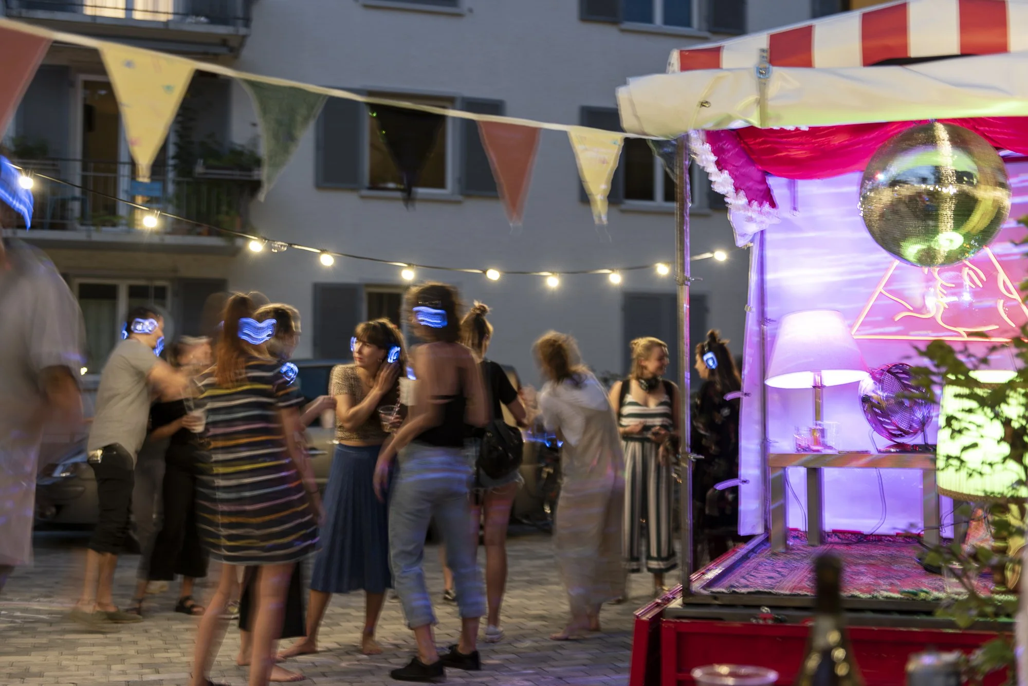 Menschen feiern auf einer Party im Freien bei Nacht, mit bunten Lichterketten und Dekorationen, einer Discokugel und einem versteckten, pink beleuchteten Bereich mit Lampen und Teppichen.