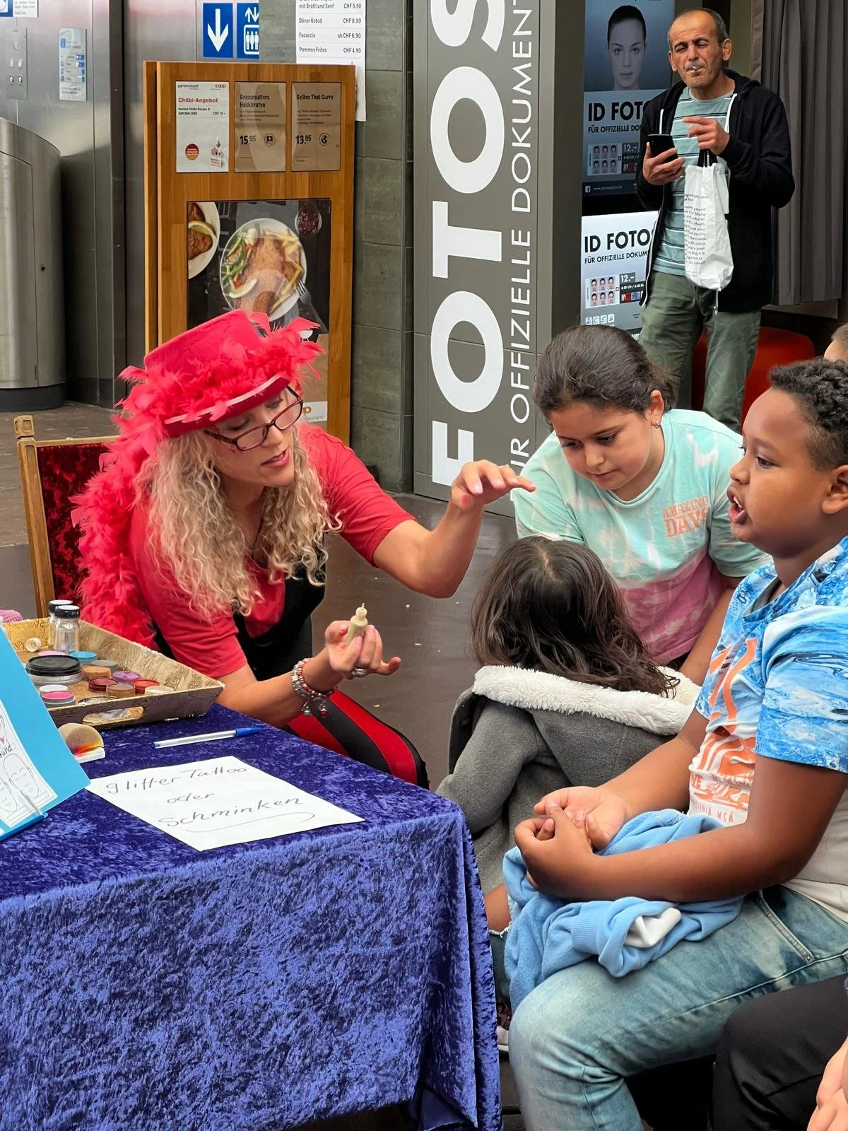 Eine Frau in einem roten Hut mit Federn zeigt Kindern bei einem Bubble-Tattoo-Workshop in einem öffentlichen Raum, mit einem Tabellenblatt, das 'Bubble Tattoo oder schminken' schreibt, und einer Palette mit verschiedenen Farben