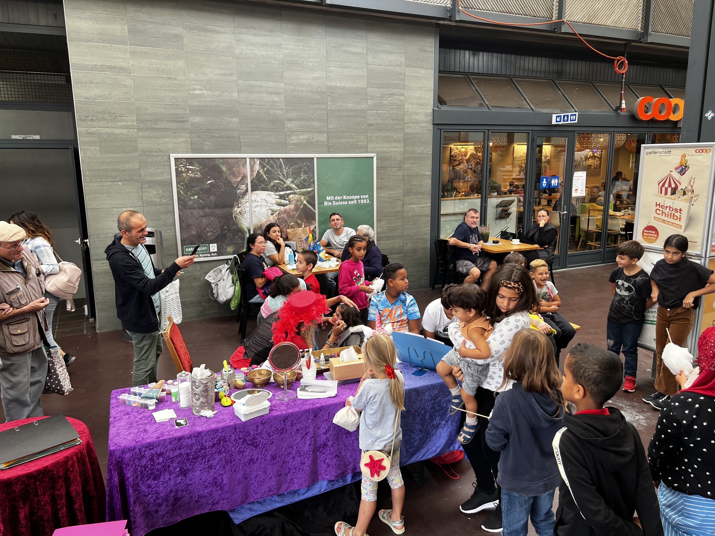 Eine Gruppe von Kindern und Erwachsenen, die sich auf einer Terrasse im Freien versammelt haben, einige sitzen an Tischen, andere stehen. Es gibt bunte Dekorationen, und in der Mitte ist ein Tisch mit verschiedenen Gegenständen. Es herrscht eine fröh