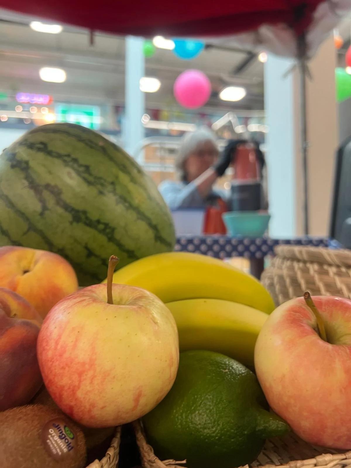 Verschiedene Früchte, darunter Äpfel, Banane, Wassermelone, Limette, und Pfirsich, in einem Korb im Supermarkt, mit einem Verkäufer im Hintergrund.