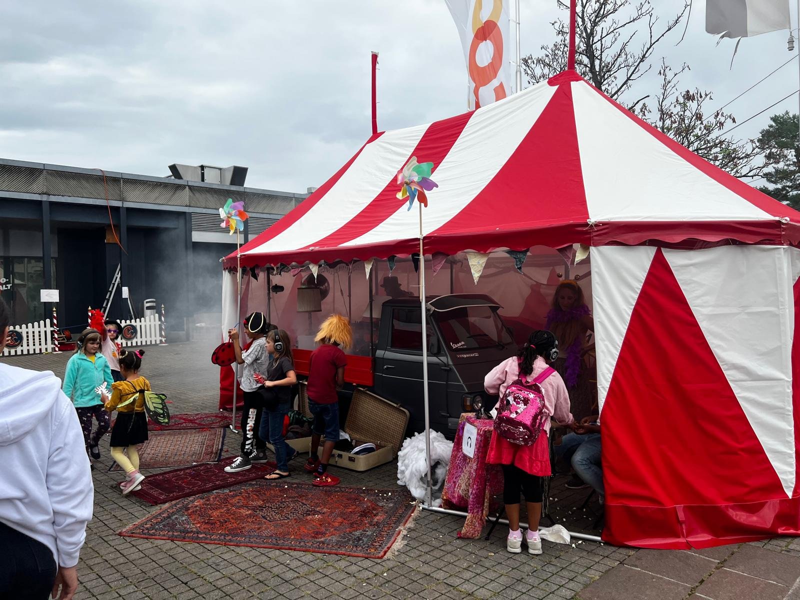 Kinder in Karnevalskostümen vor einem roten und weißen Zirkuszelt, das eine Spielstation mit einer kleinen Kasse enthält.