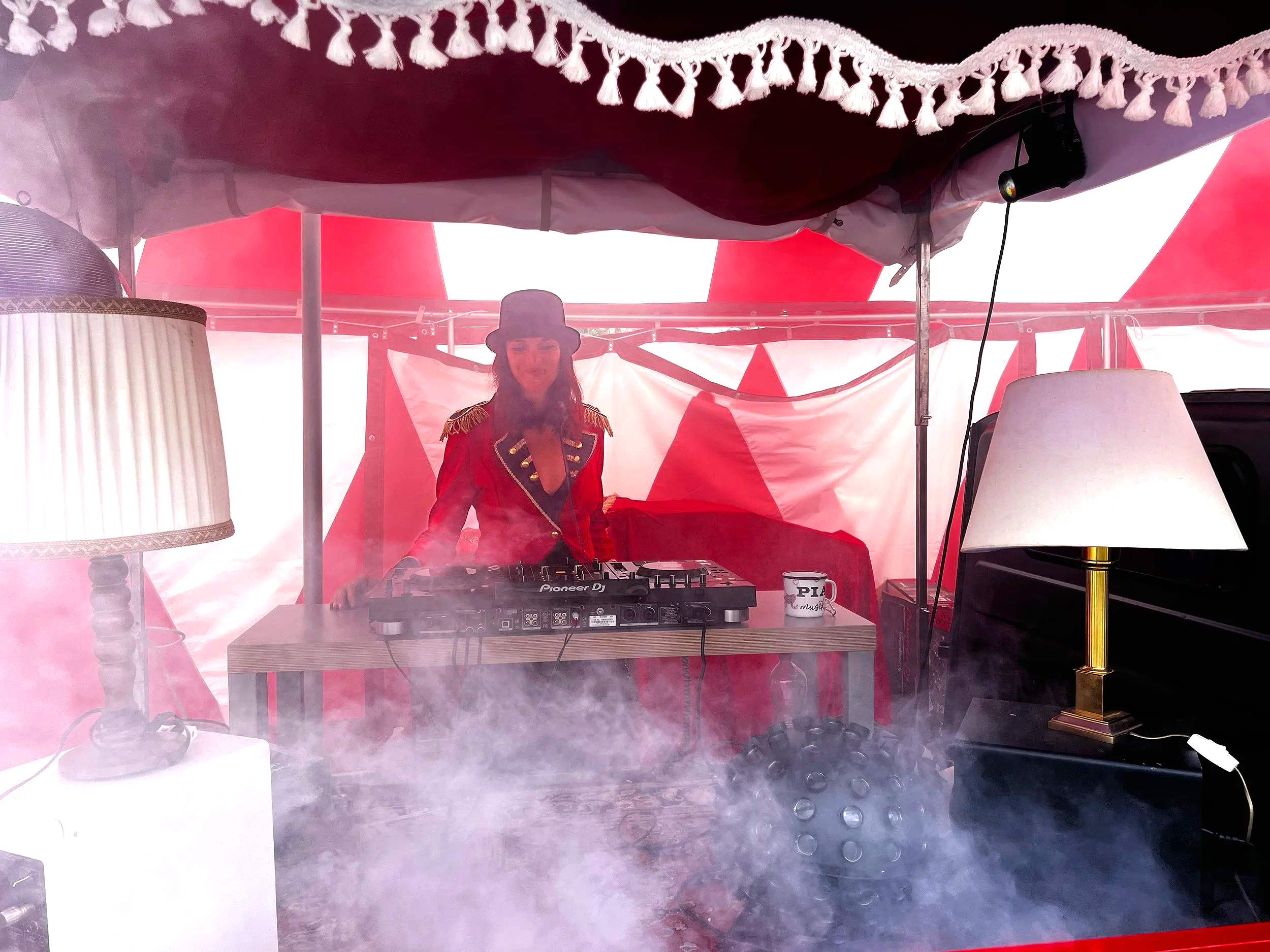 Eine Frau in einem roten Anzug mit Schulterepauletten und Hut, die als DJ an einem Mischpult hinter einem Zelt auftritt, umgeben von Lampen und Rauch.