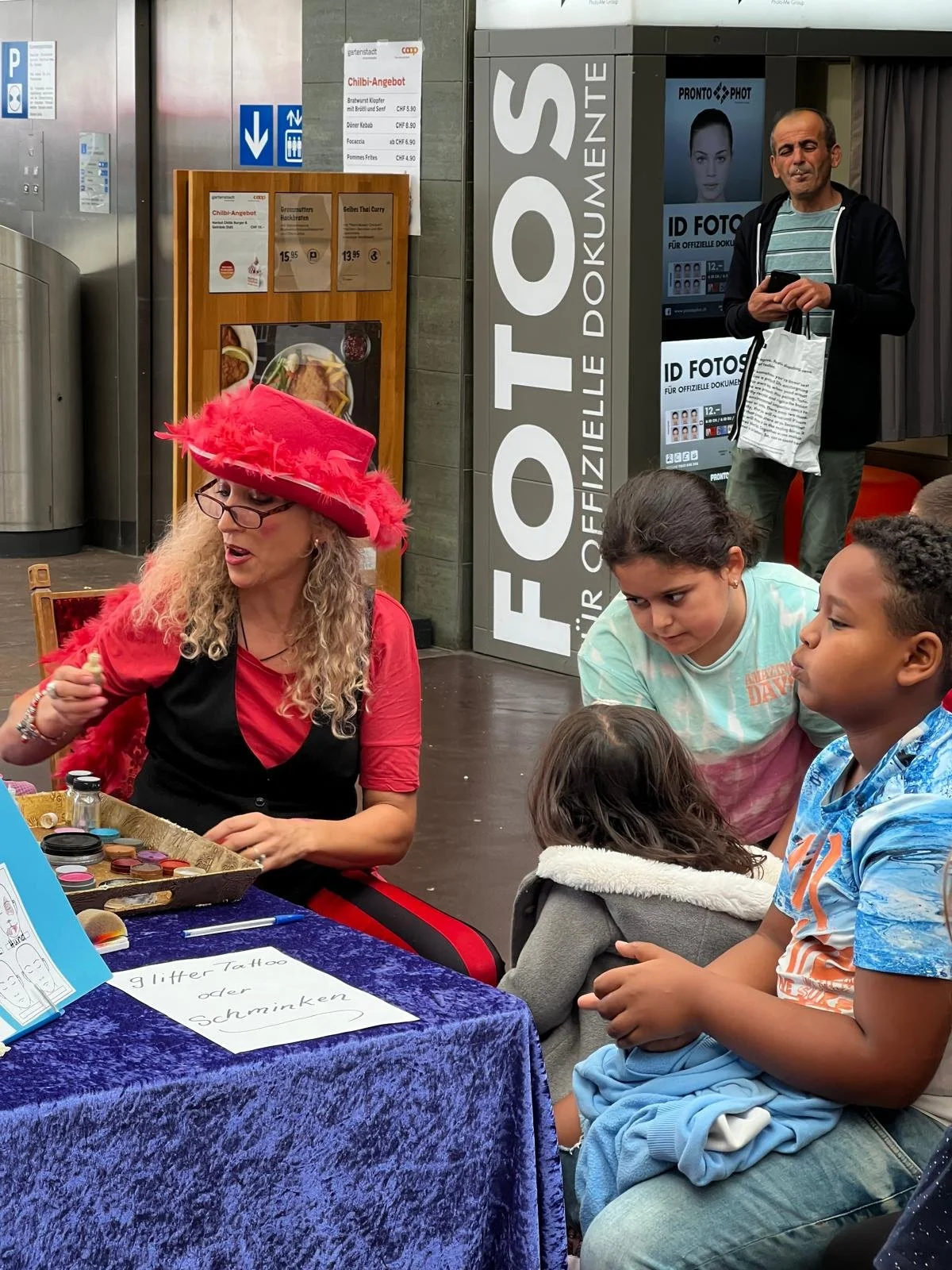 Eine Frau in rotem Kleid und pinkem Hut, die Tätowierungen oder Schminke illustriert, sitzt an einem Tisch mit Kindern, die interessiert zusehen. Es gibt einen signierten Schilder mit der Aufschrift 'Glitter Tattoo oder Schminken' auf dem Tisch. Im H