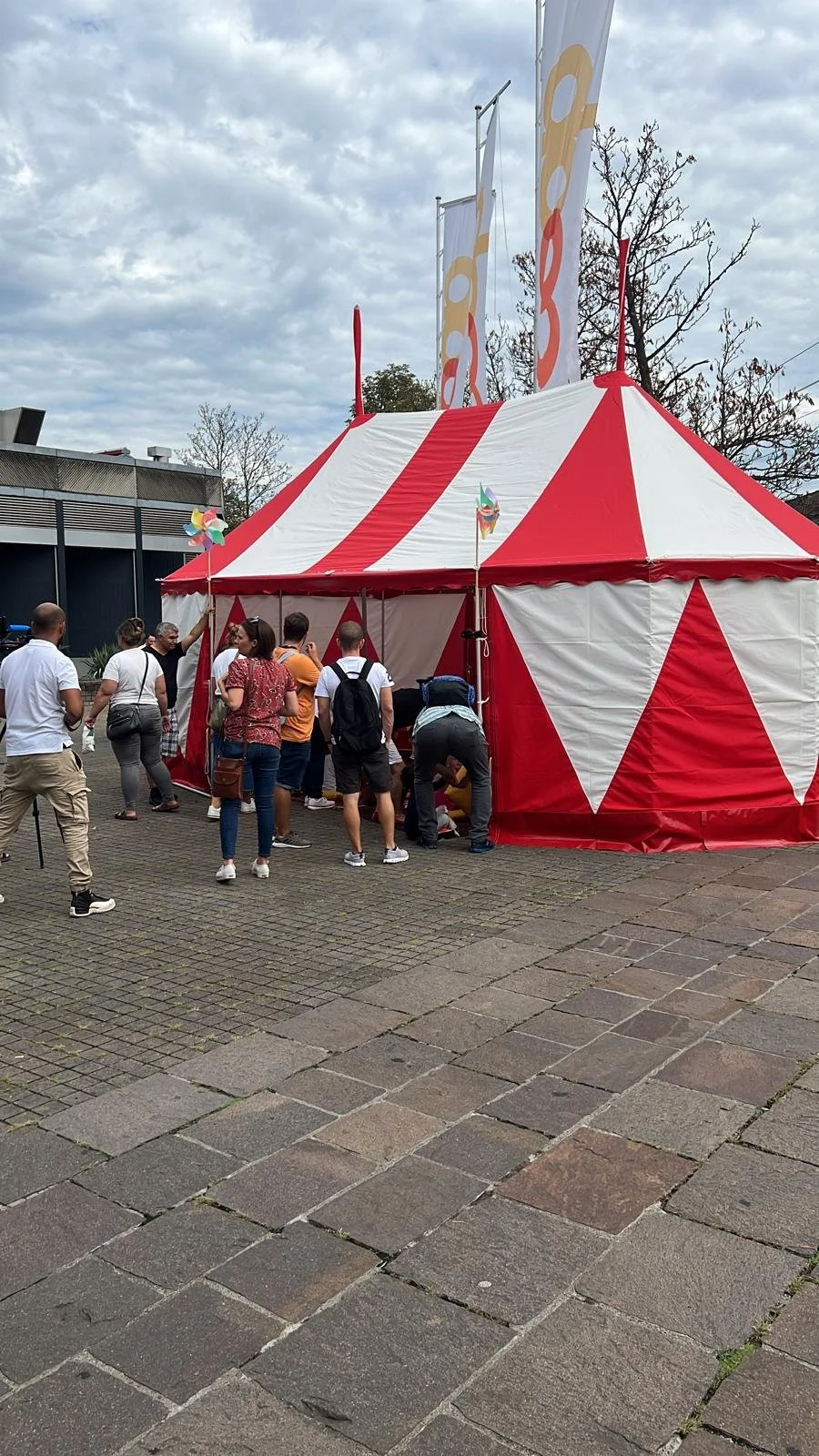 Menschen vor einem rot-weiß gestreiften Zirkuszelt an einem Tag mit bewölktem Himmel.