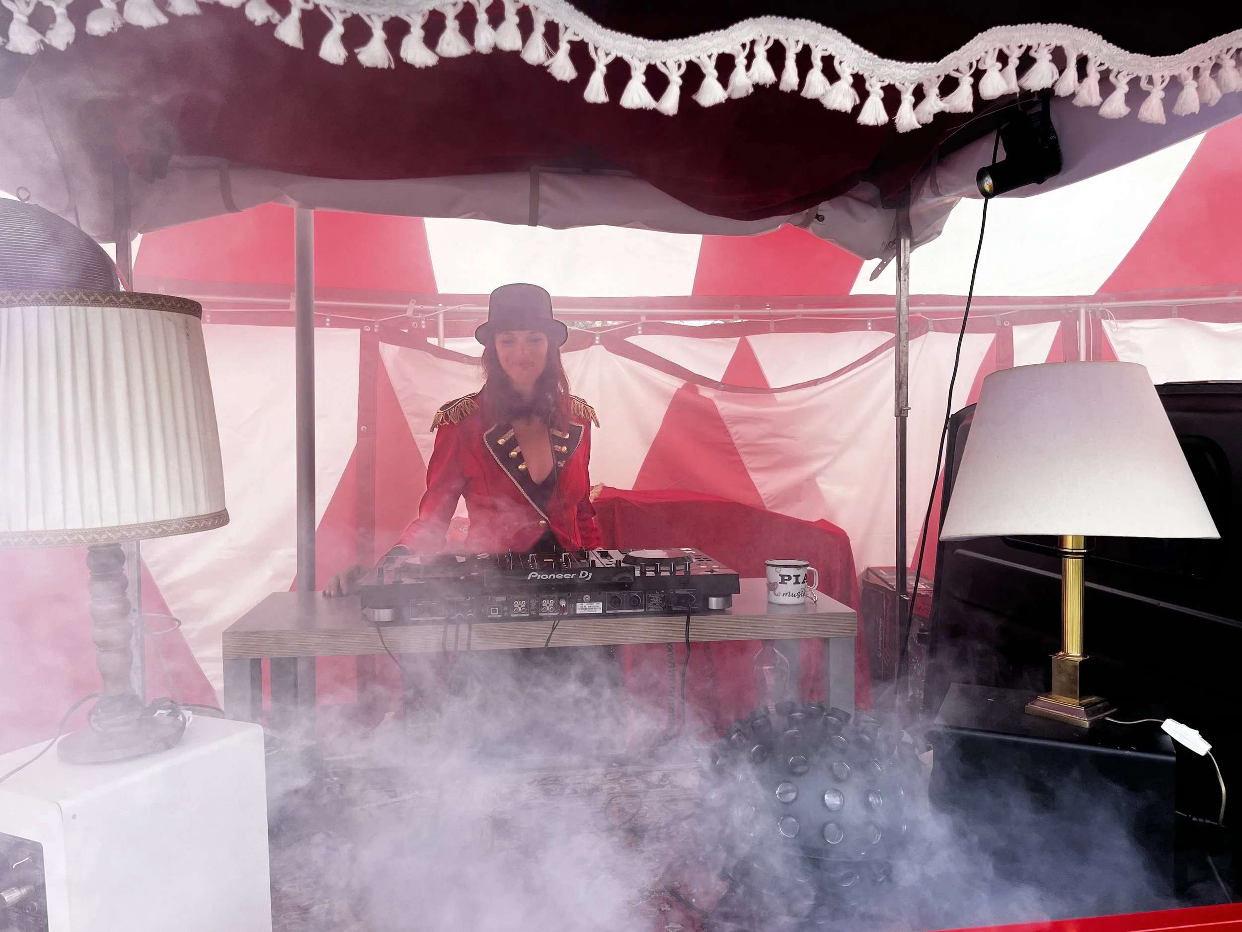 Eine Frau in rotem Jacke und Zylinderkopfstück steht hinter einem DJ-Mischpult in einer Manege, umgeben von Zirkusstimmung mit Rauch, Lampen und Zirkuszelt. 