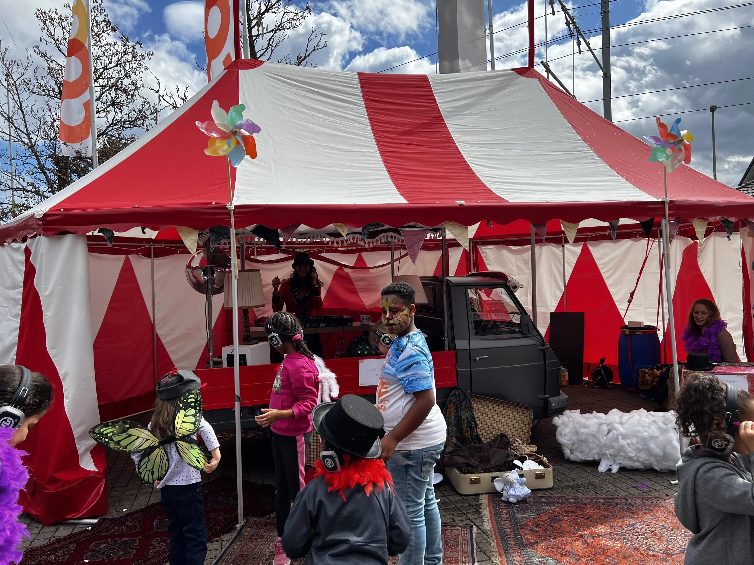 Kinder mit Kopfhörern, die auf einer Zirkus- oder Festzelt-Bühne tanzen, umgeben von dekorativem Ballon und künstlerischer Requisite, bei einem Fest im Freien.