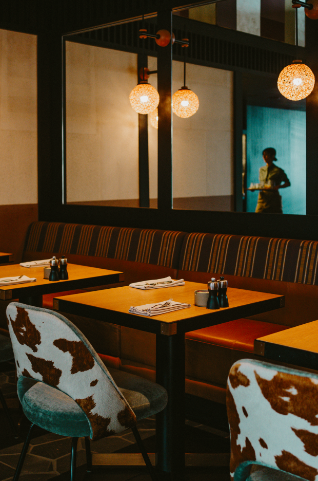 Empty restaurant with wooden tables set with napkins, utensils, and salt and pepper shakers. Cow-print and striped upholstered chairs. Warm lighting from hanging spherical lamps. A server is visible through a mirror on the wall.