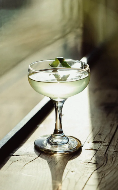 Cocktail glass with lime wedge, on a wooden surface near a window.