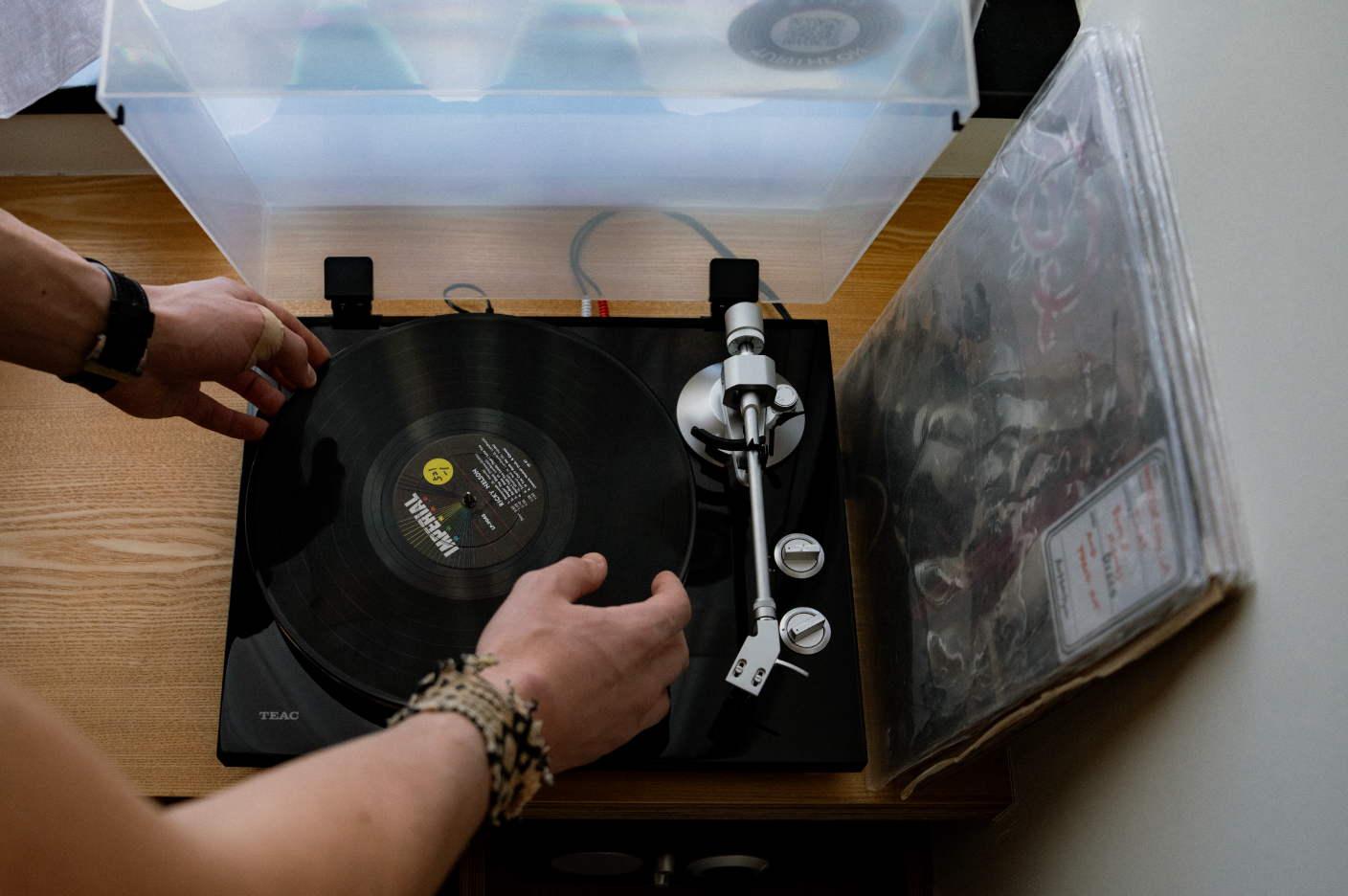 Person handling a vinyl record on a turntable, placed on a wooden surface with a clear plastic cover, and a vinyl record box on the right.