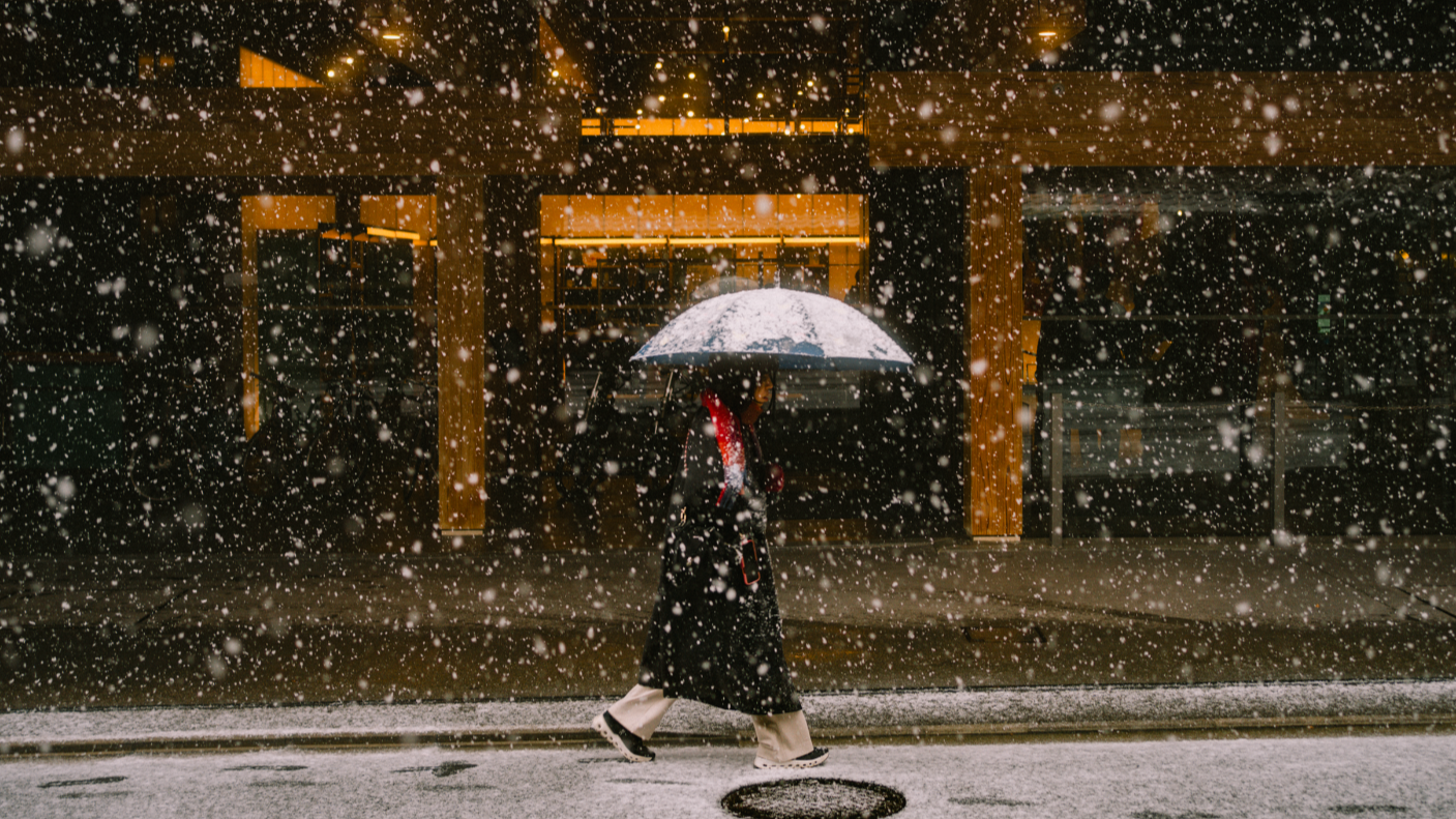 A passerby walks with an umbrella and a red scarf in front of a hotel lobby during a snowy day
