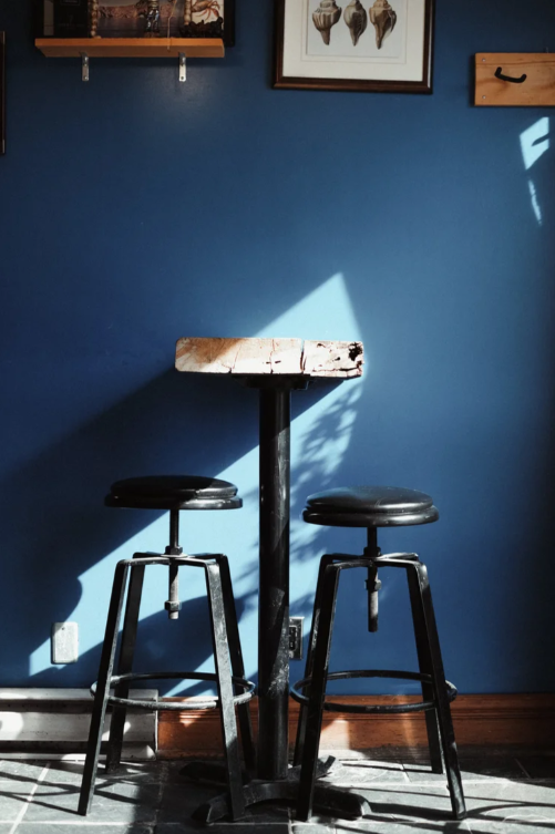 A small wooden table with two black bar stools against a blue wall, with sunlight creating shadows on the wall and floor.