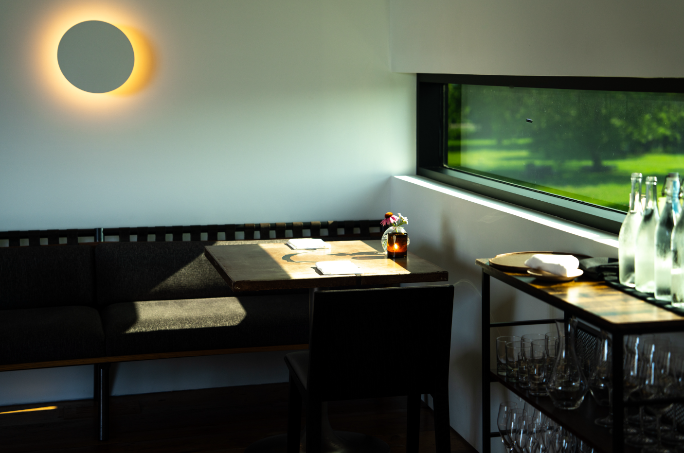 A cozy dining area with a dark couch, a wooden table with a vase of flowers, sunlight streaming through a large window, and a shelf with glass bottles and glasses.