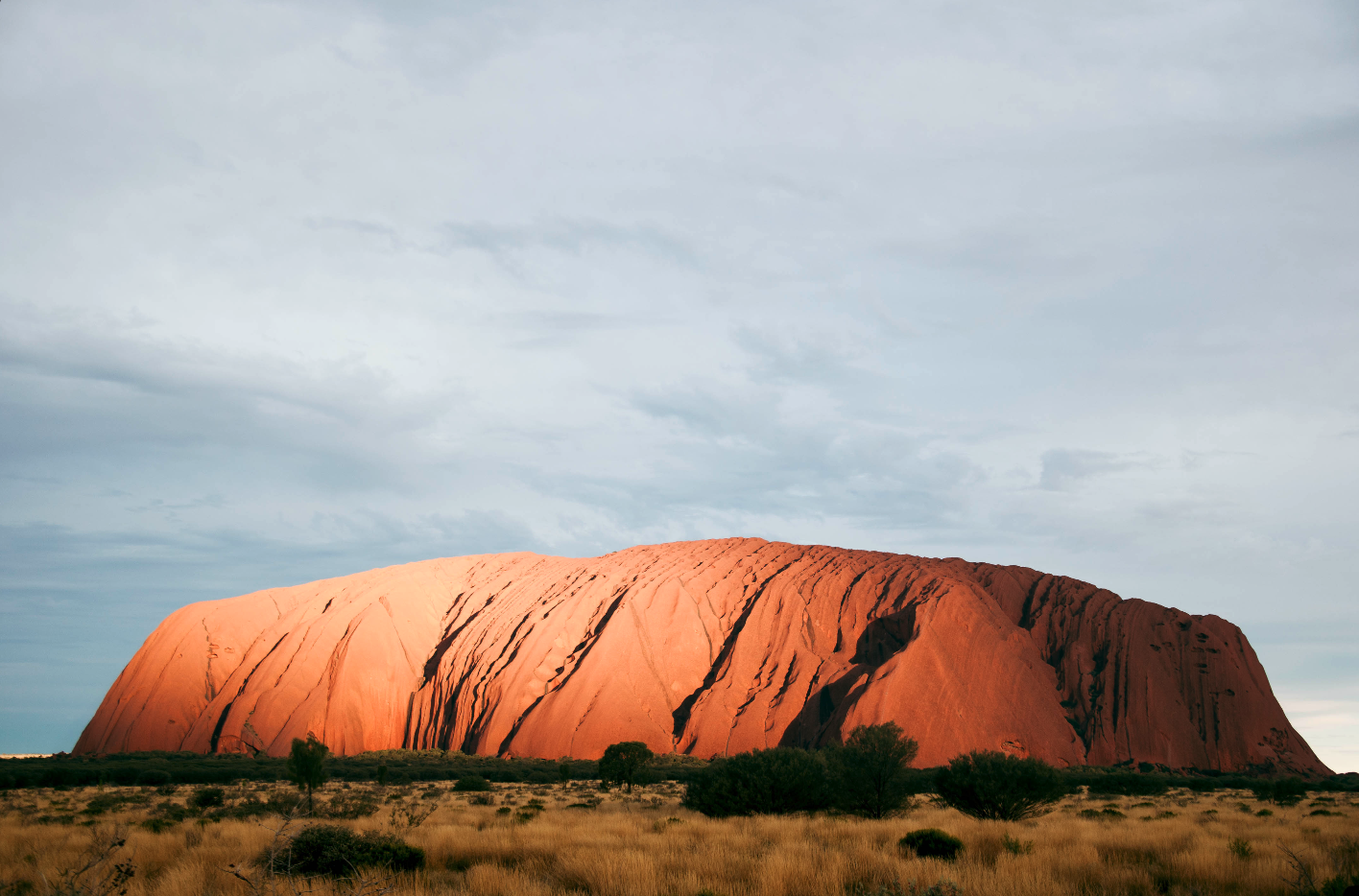 A large, reddish-brown sandstone formation known as Uluru or Ayers Rock in the Australian outback, with sparse trees and grass in the foreground and a cloudy sky above.