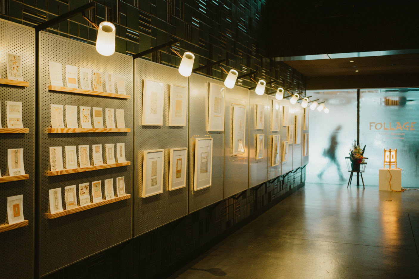 An art gallery features framed artwork on a wall with a perforated metal backdrop. Overhead, multiple white cylindrical lights illuminate the display. To the right, a frosted glass door has the word 'FOLIAGE' in large letters, and a silhouette of a p