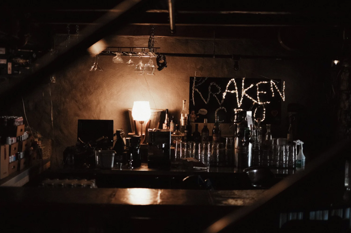 Dimly lit bar with glasses and bottles, illuminated by a small lamp, with a sign that reads 'KRAKEN' in lights.