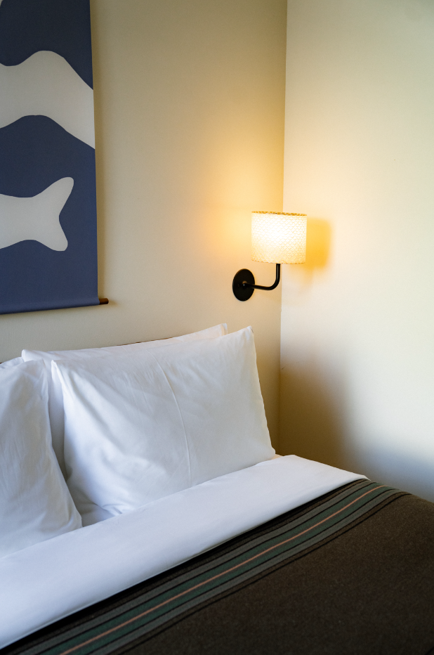 Part of a bedroom with a bed, white pillows, a wall-mounted lamp, and a blue and white abstract wall hanging.
