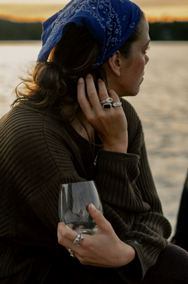 A woman with brown hair, wearing a blue bandana and a brown sweater, sitting outdoors by a body of water at sunset, holding a glass of wine.