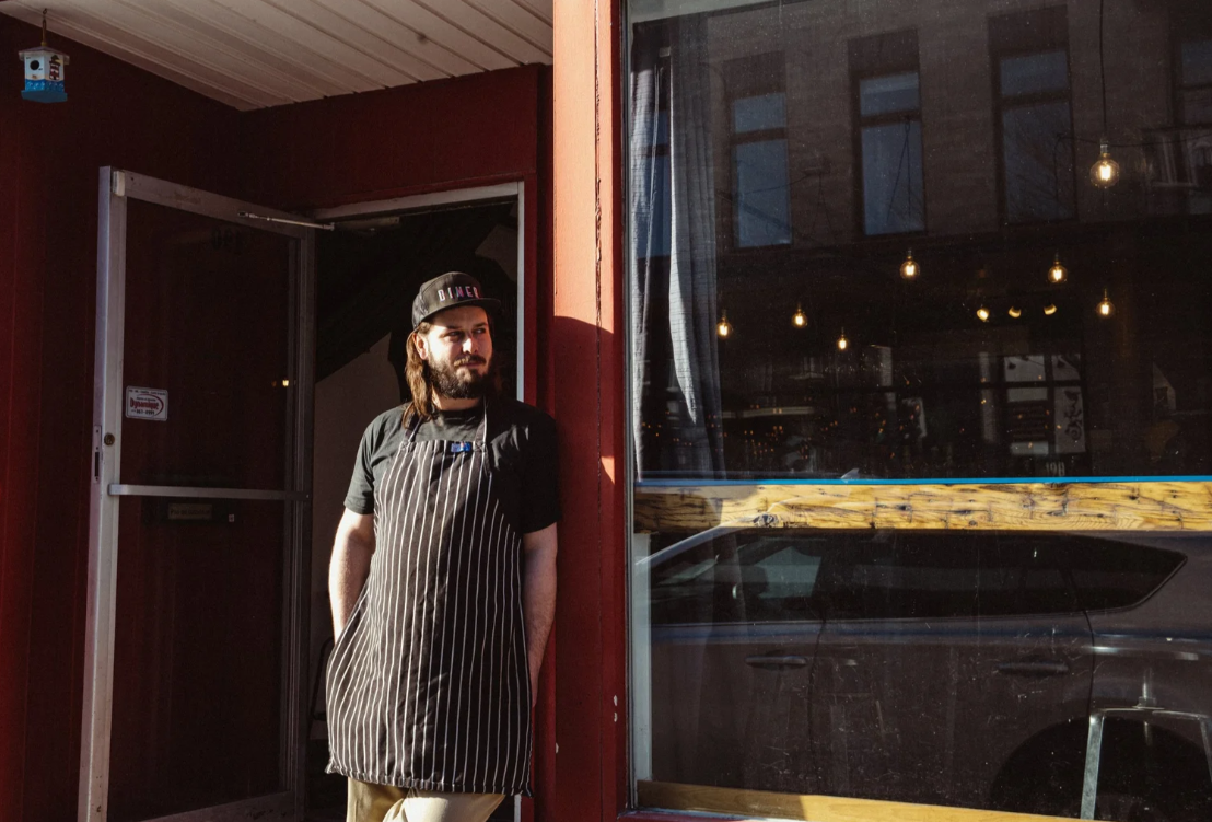 A young man with a beard, wearing a baseball cap and striped apron, stands outside a restaurant, looking to the side and leaning against the red exterior wall.